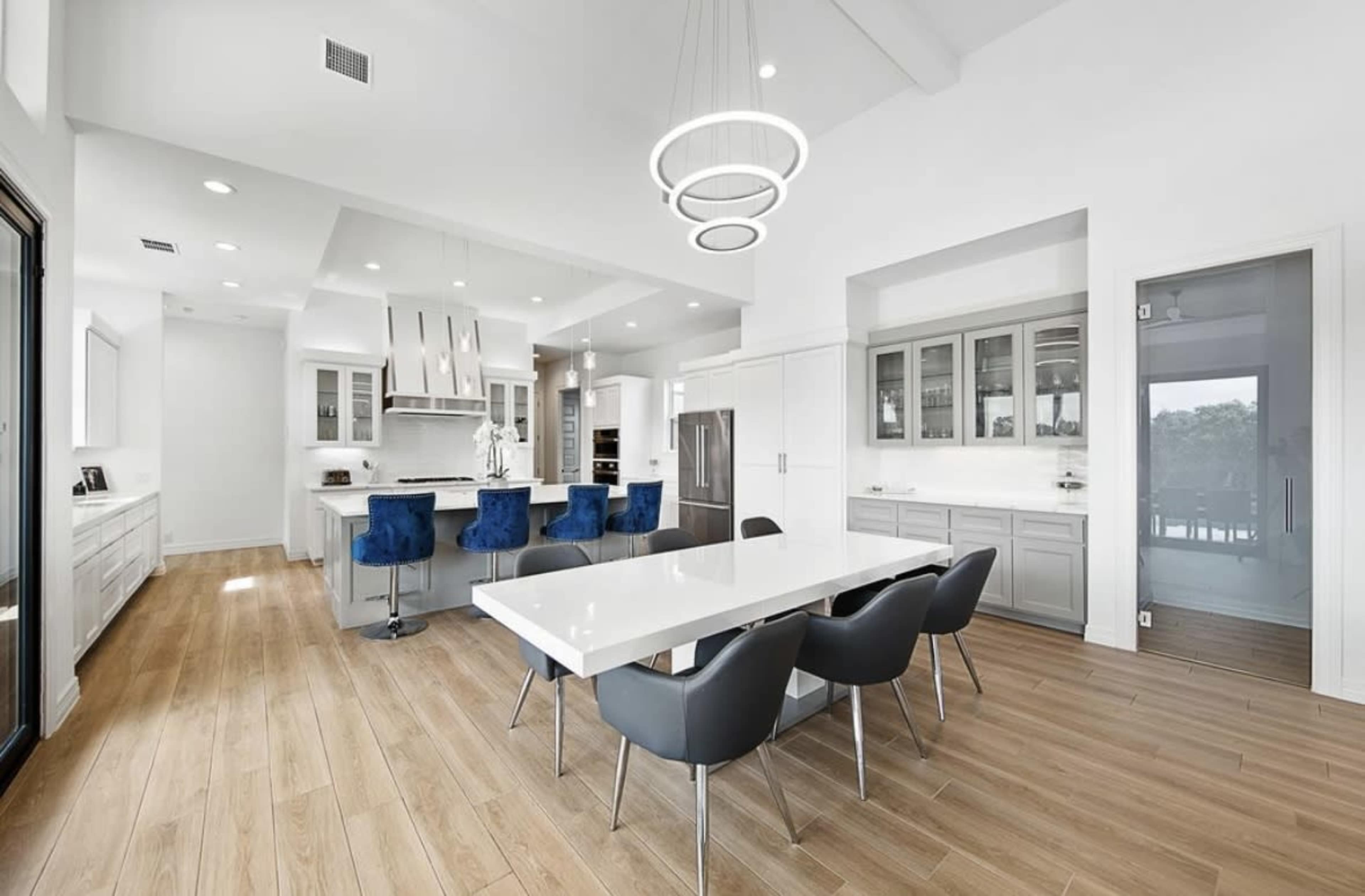 The image shows a modern kitchen and dining area featuring a long white dining table, gray chairs, and blue upholstered bar stools around an island, with large windows allowing natural light to fill the space.