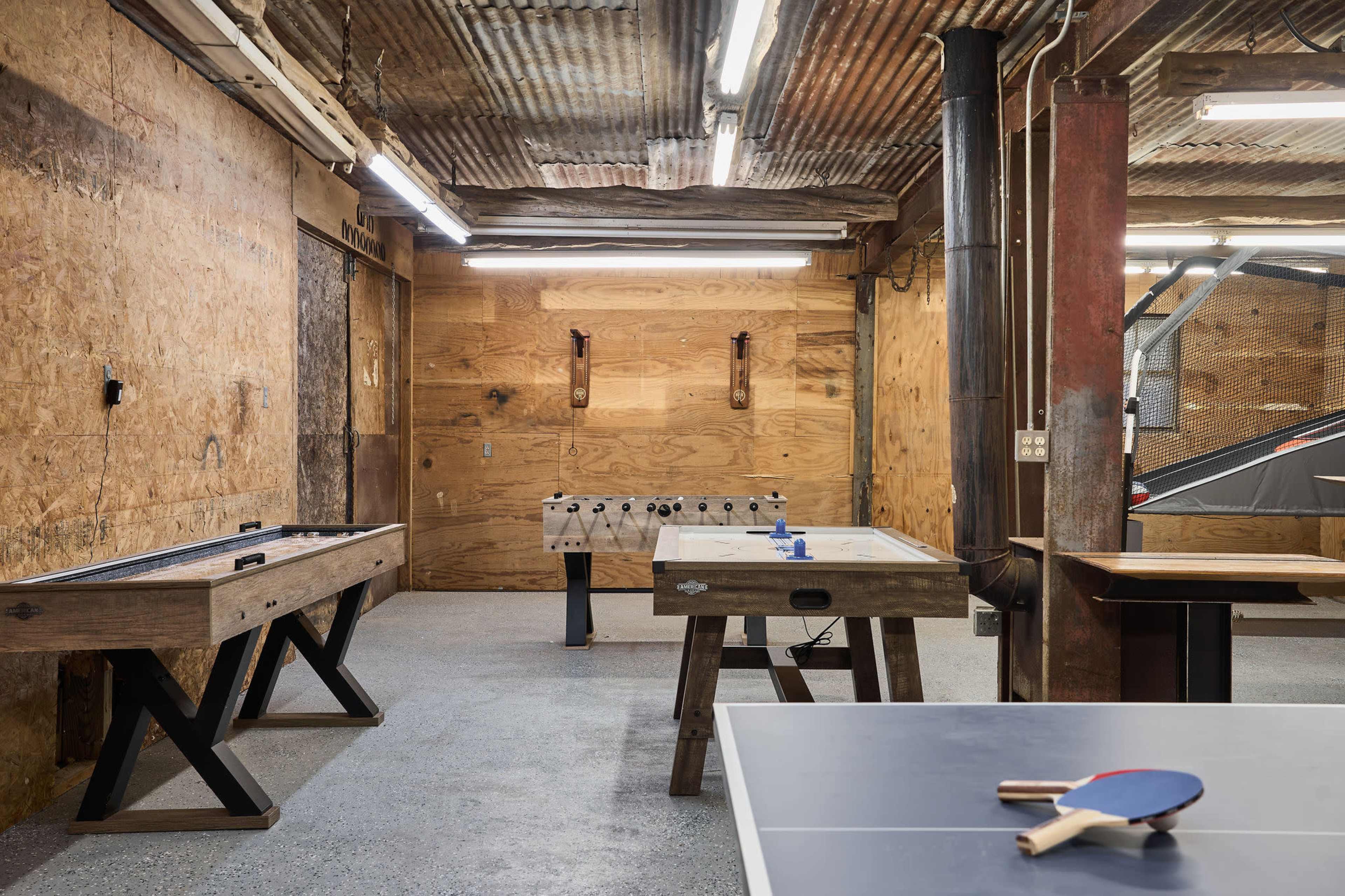 The image shows a multi-purpose game room with a ping pong table, foosball table, and air hockey table set within a rustic, industrial-style interior featuring wooden walls and exposed lighting.