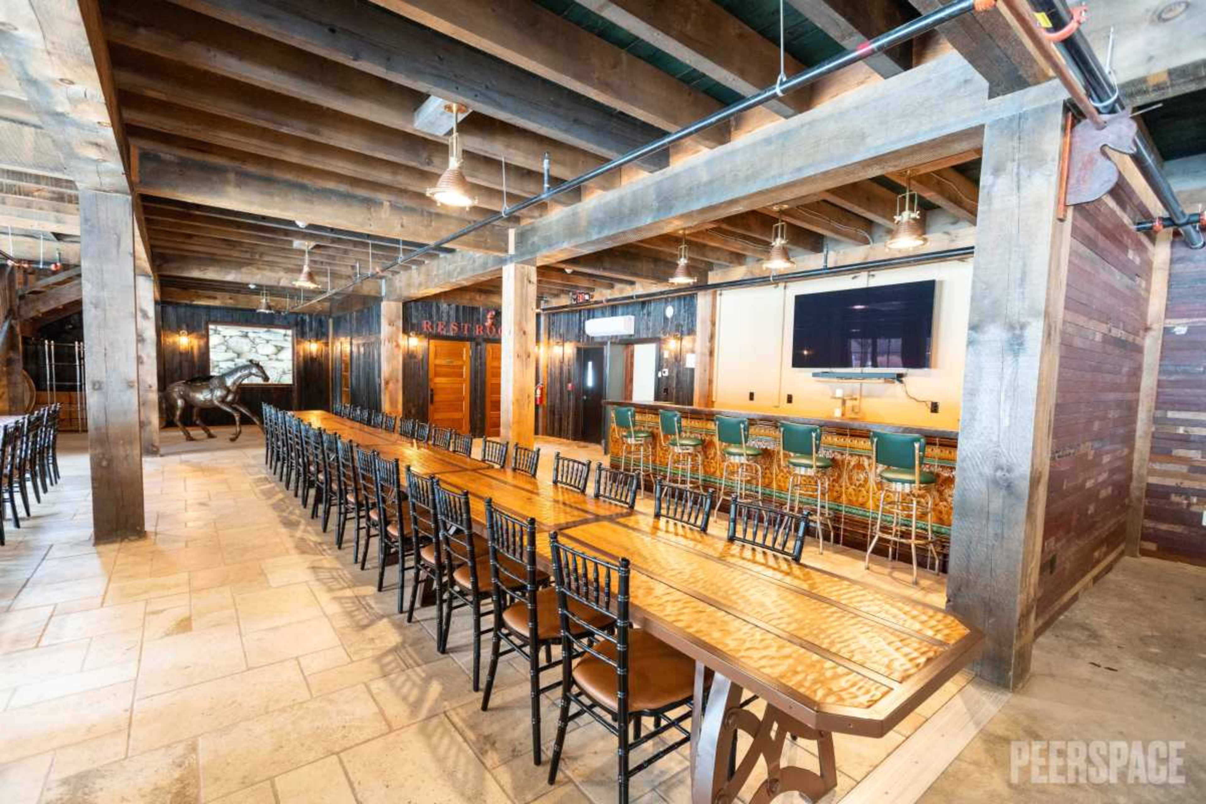 The image shows a spacious dining area with long wooden tables and a bar, featuring exposed wooden beams and stone accents.