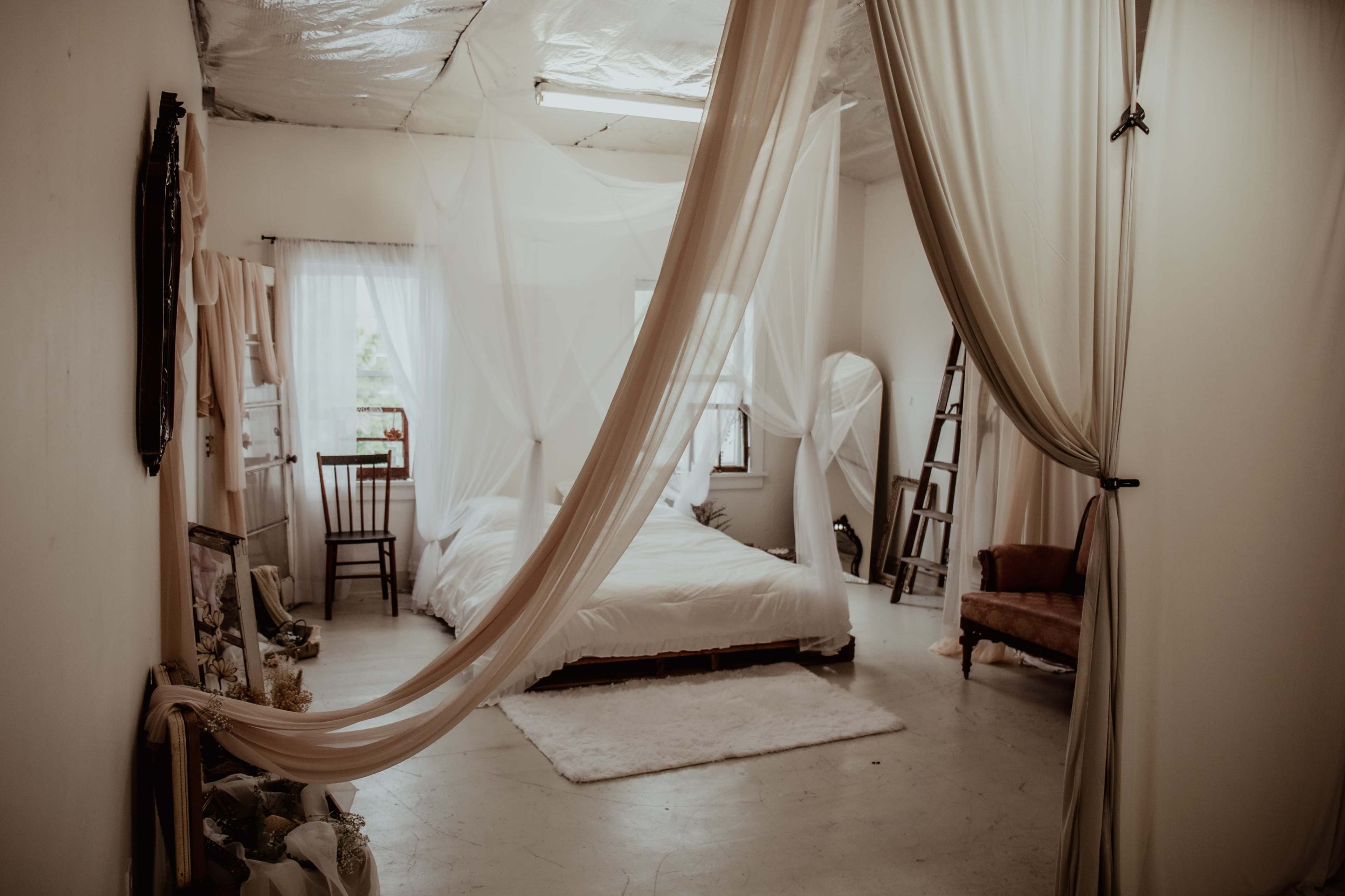 A minimalistic bedroom with a bed draped in white fabric, a chair, and a ladder against the wall.