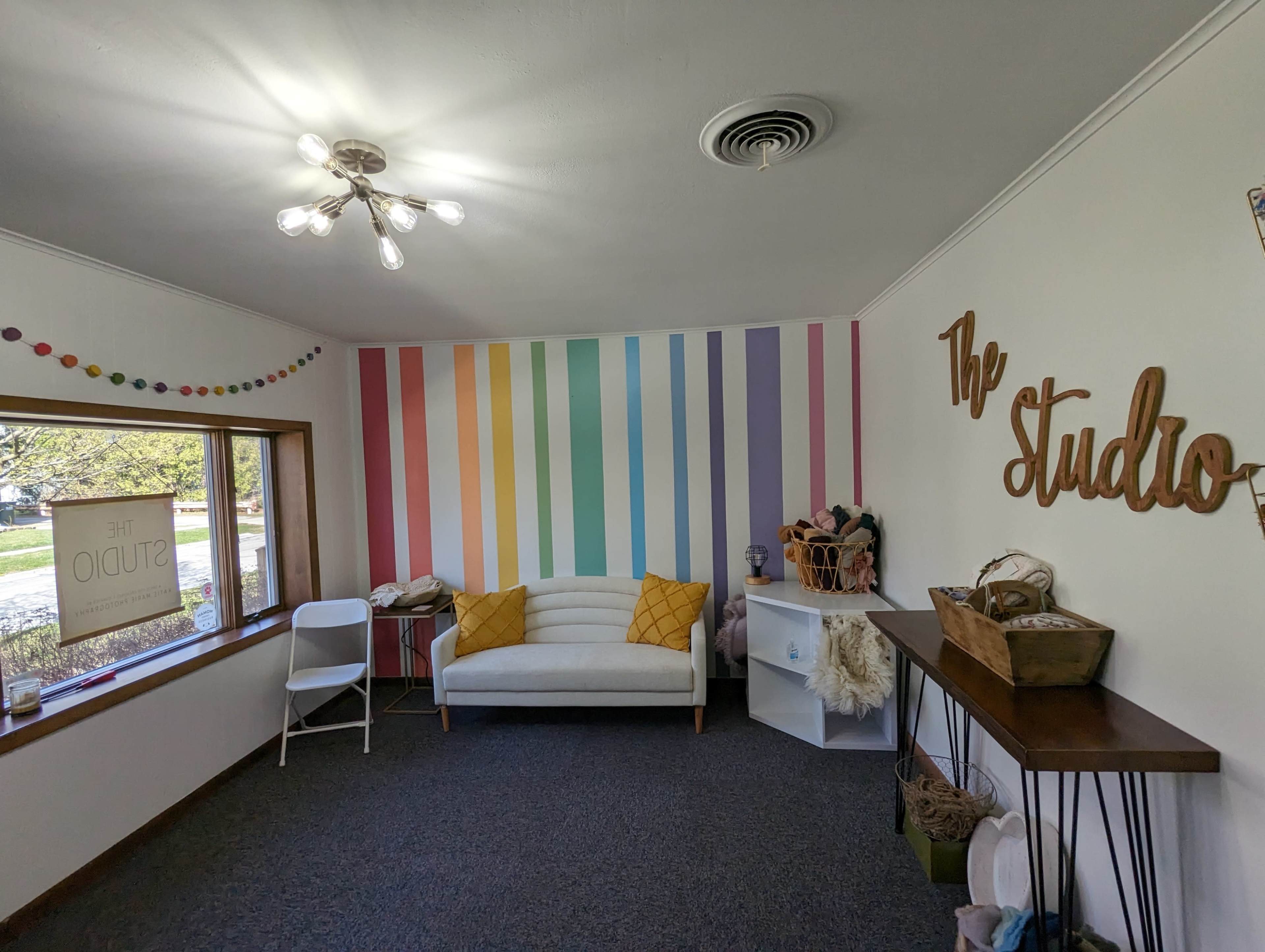 A bright room features a white couch with yellow cushions, colorful striped walls, and a decorative wooden sign that reads "The Studio."