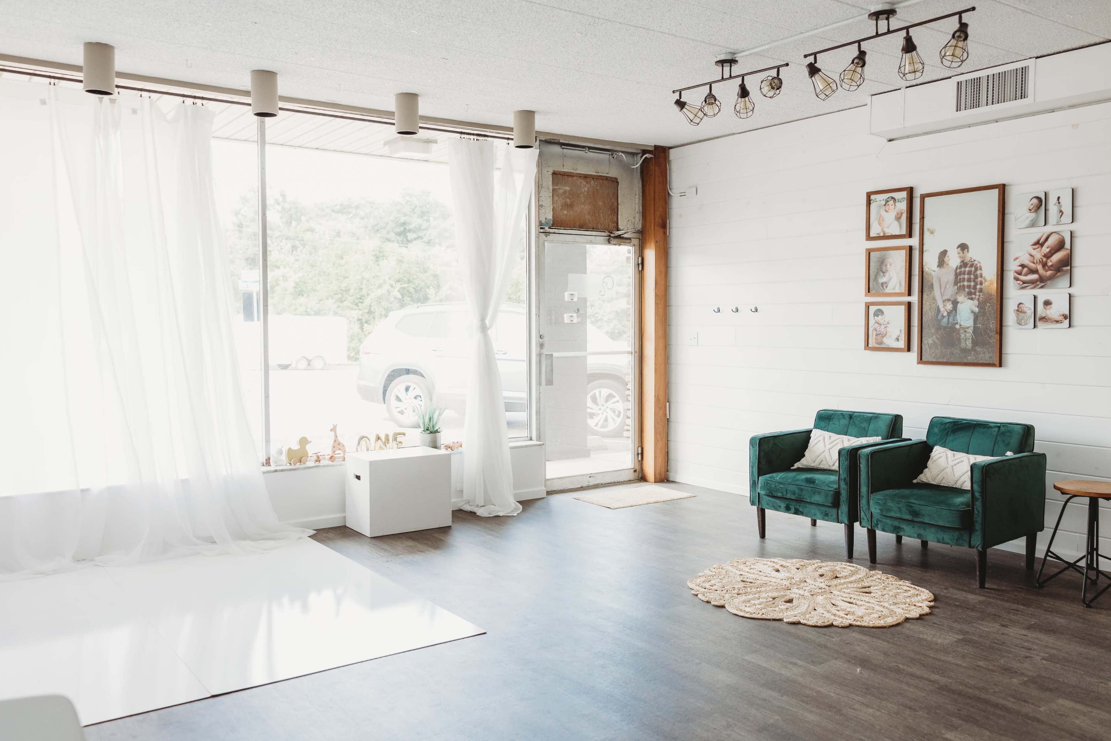 A bright, minimalist interior space with two green armchairs, a white backdrop, and large windows allowing natural light to enter.