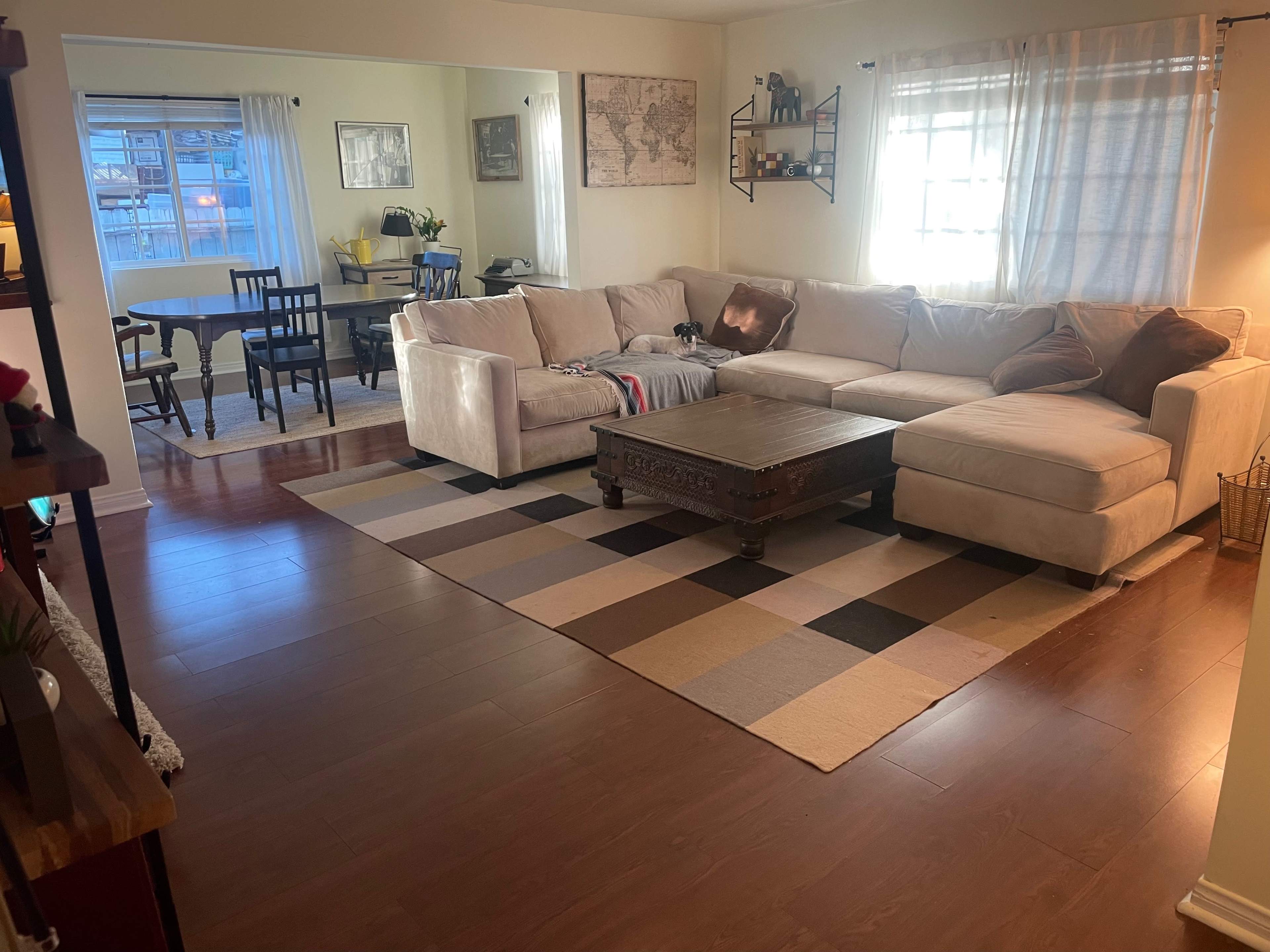 The image shows a living room with a large beige sectional sofa, a dark wooden coffee table, and a patterned area rug, along with a dining area visible in the background.