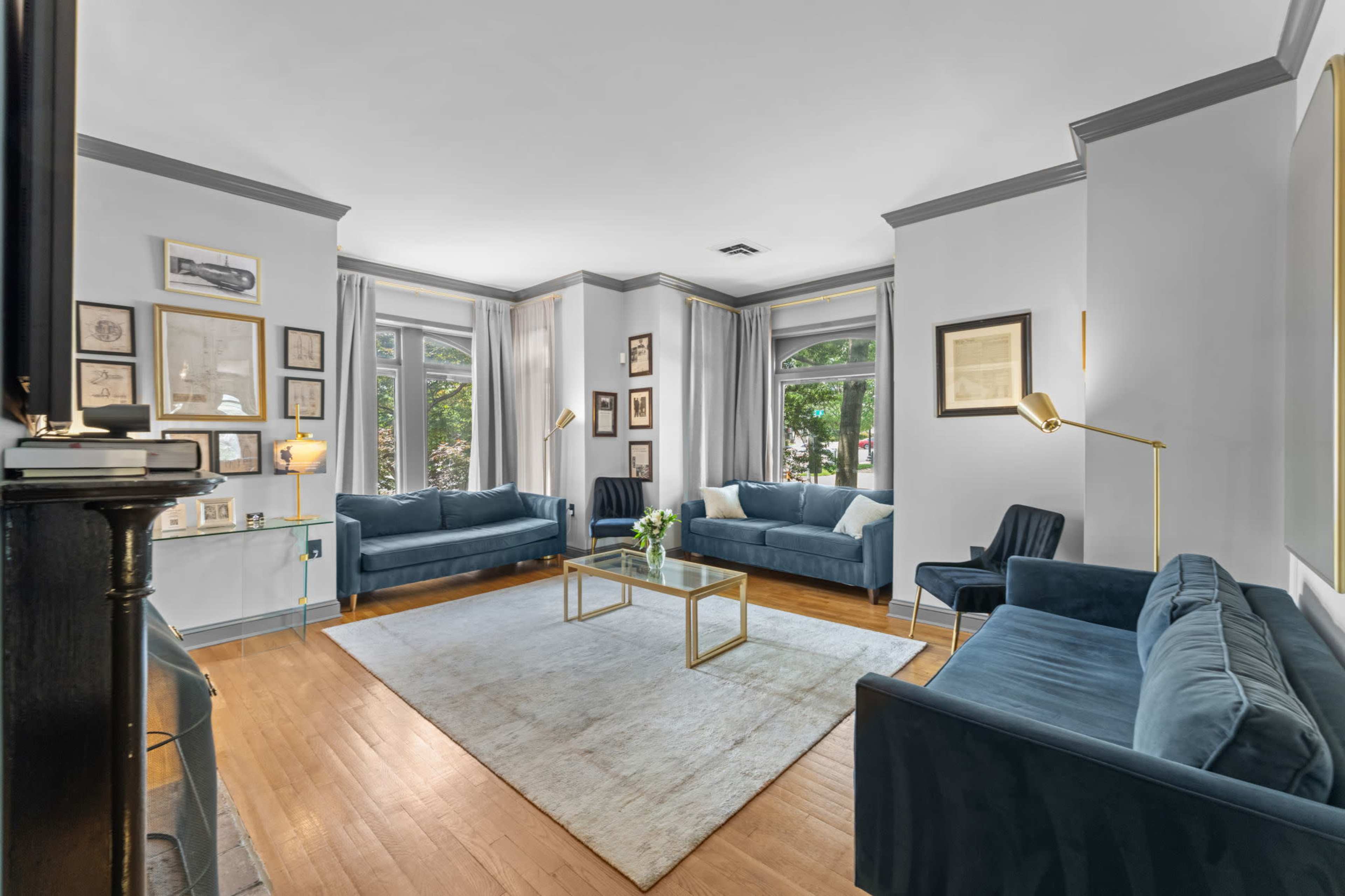 The living room features two blue sofas and a black armchair arranged around a glass coffee table, with framed artwork on the walls and large windows allowing natural light to enter.
