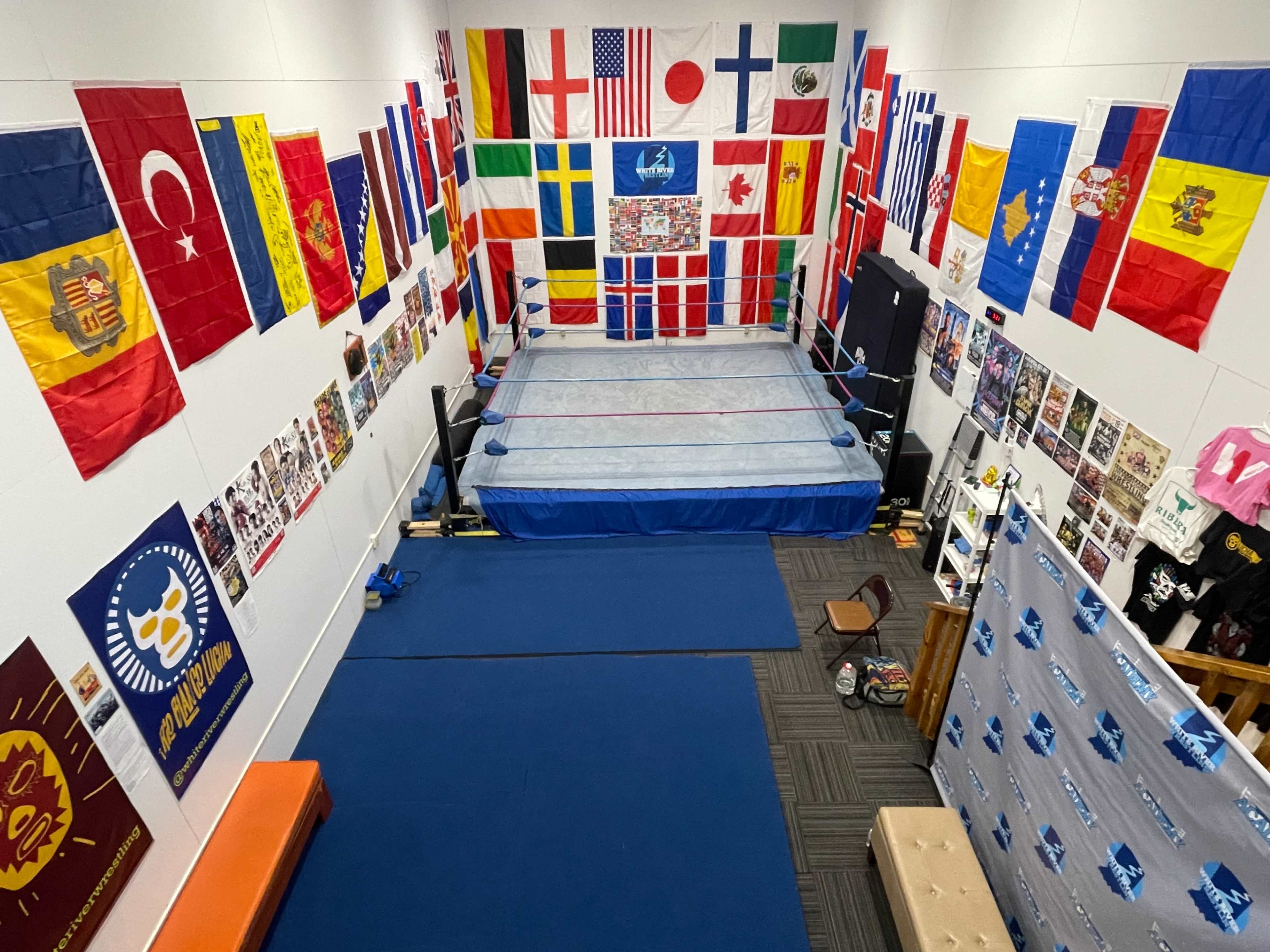 The image shows an indoor wrestling ring surrounded by various national flags and posters on the walls.