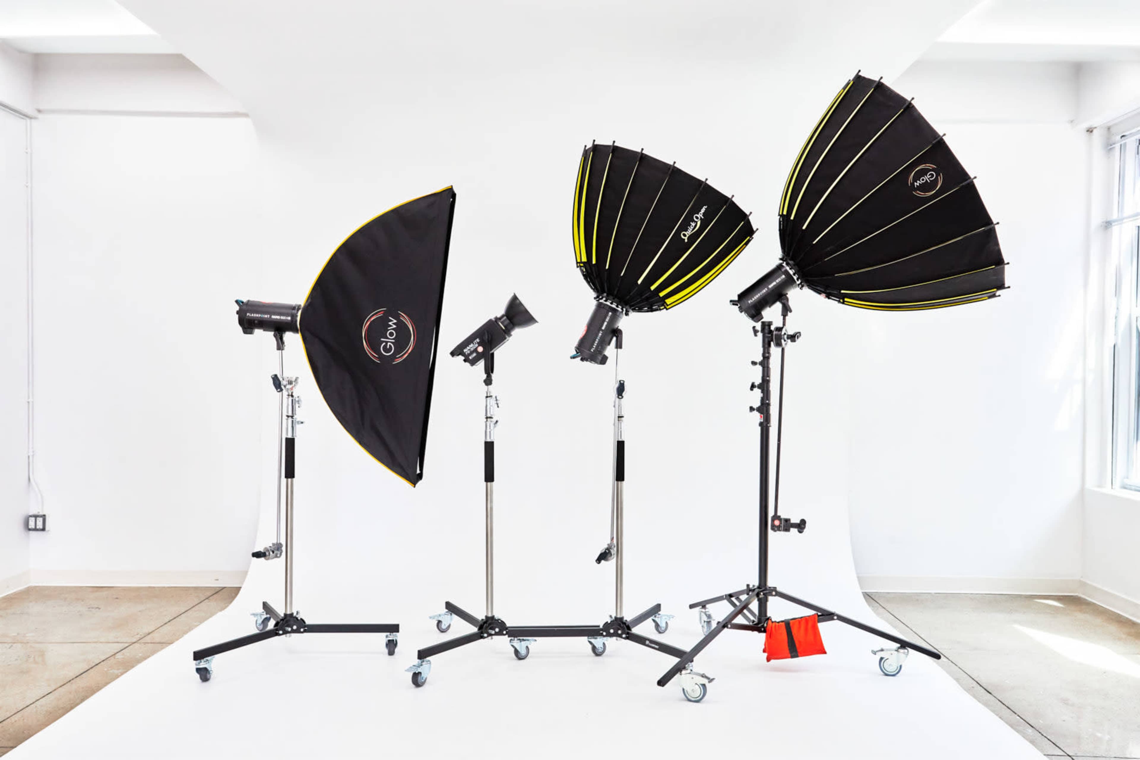 The image shows a row of photography lights positioned on stands against a white backdrop in a studio setting.