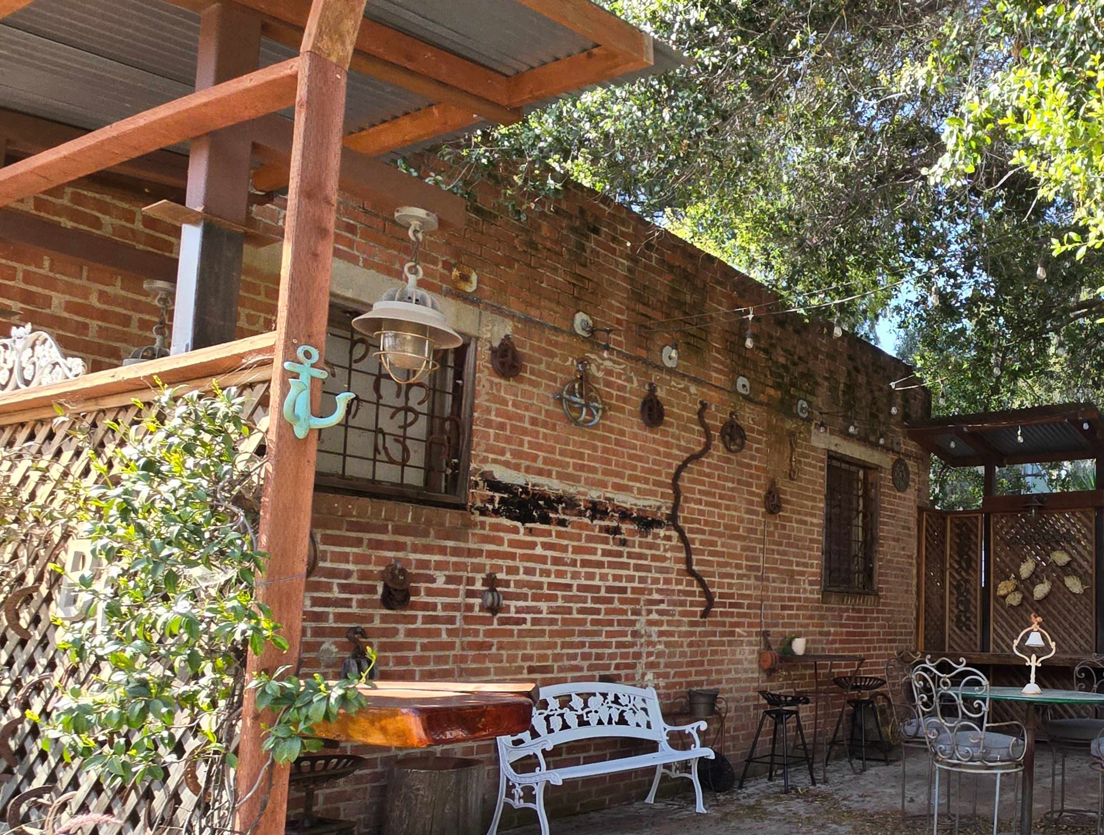 A patio area with a brick wall adorned with various decorations, including metal anchors and a winding vine, next to outdoor furniture arranged under a shaded roof.