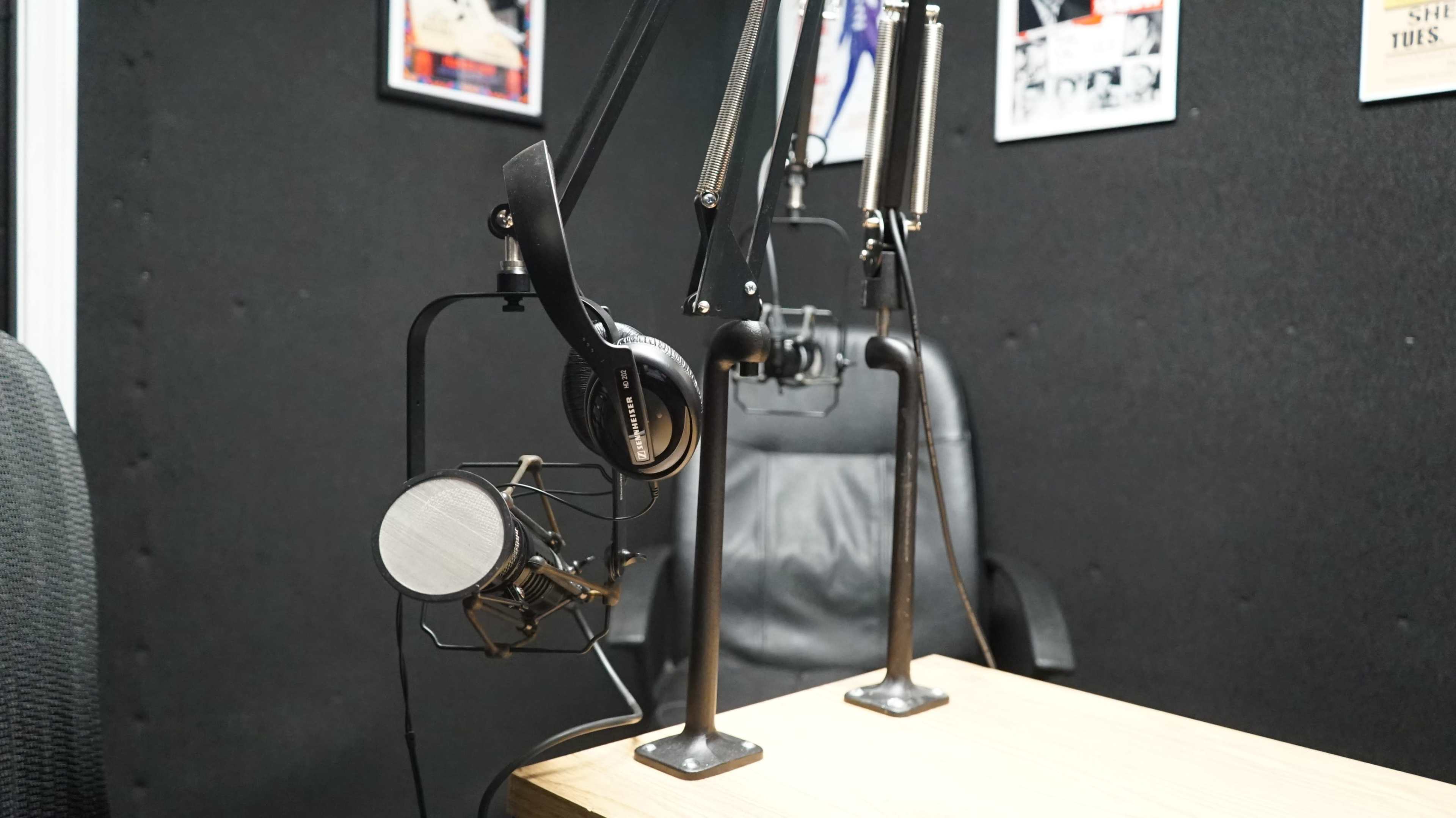 The image shows a radio studio setup with a microphone on a stand, headphones hanging nearby, and a chair against a black wall adorned with posters.