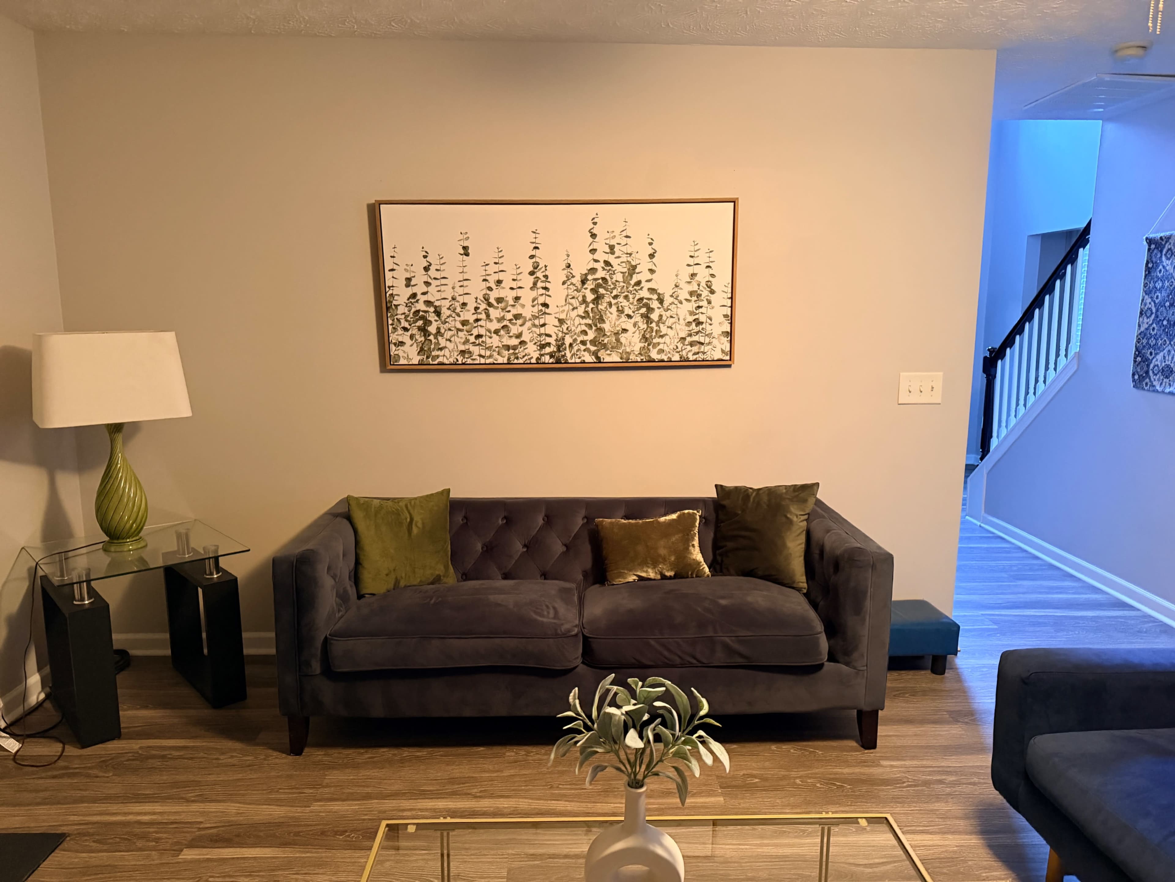 A modern living room features a dark sofa with decorative pillows, a glass coffee table, a lamp, and a piece of artwork on the wall, with a staircase visible in the background.