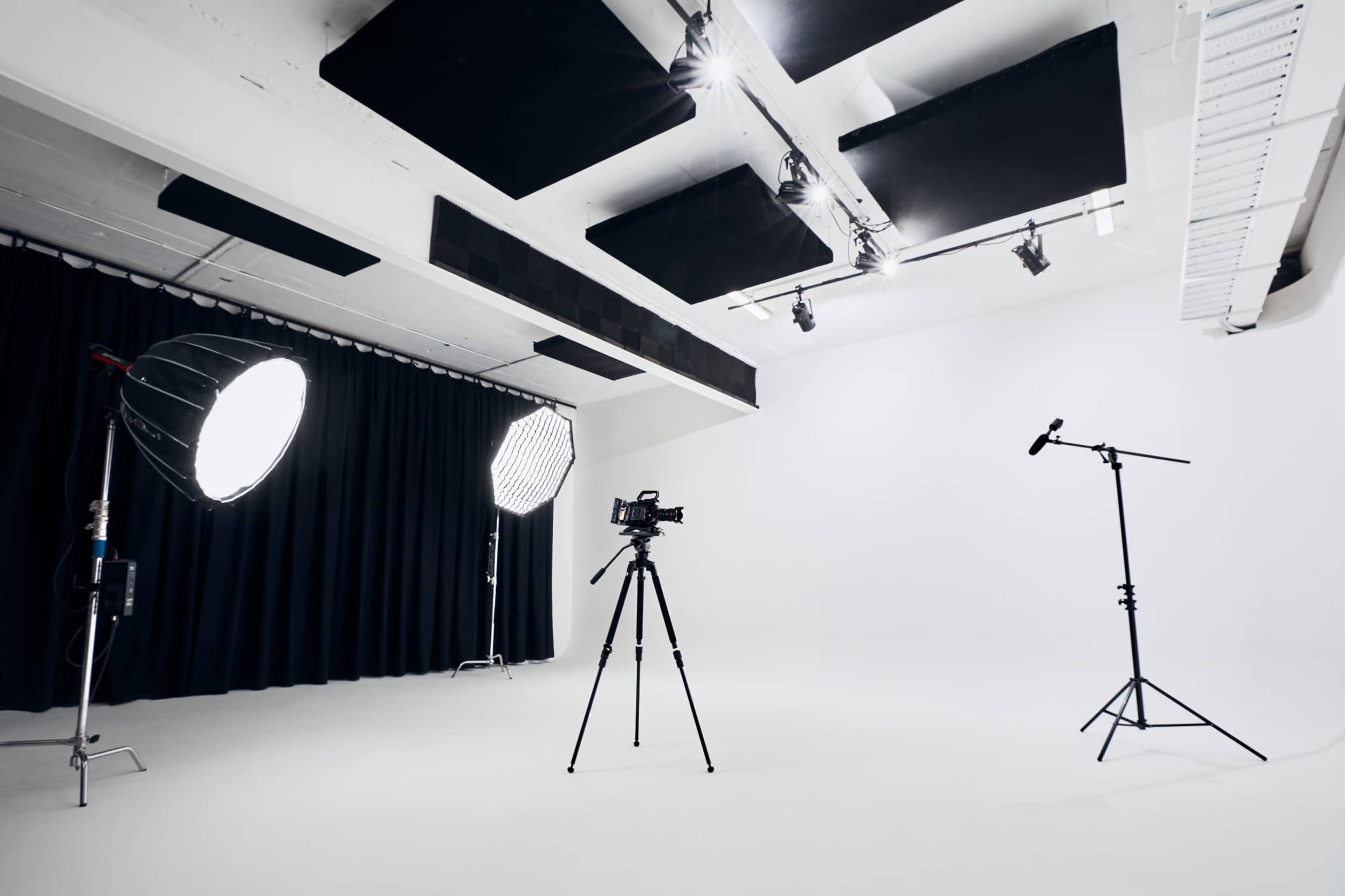 The image shows a photography studio equipped with a camera on a tripod, softbox lights, and a microphone stand against a plain backdrop.