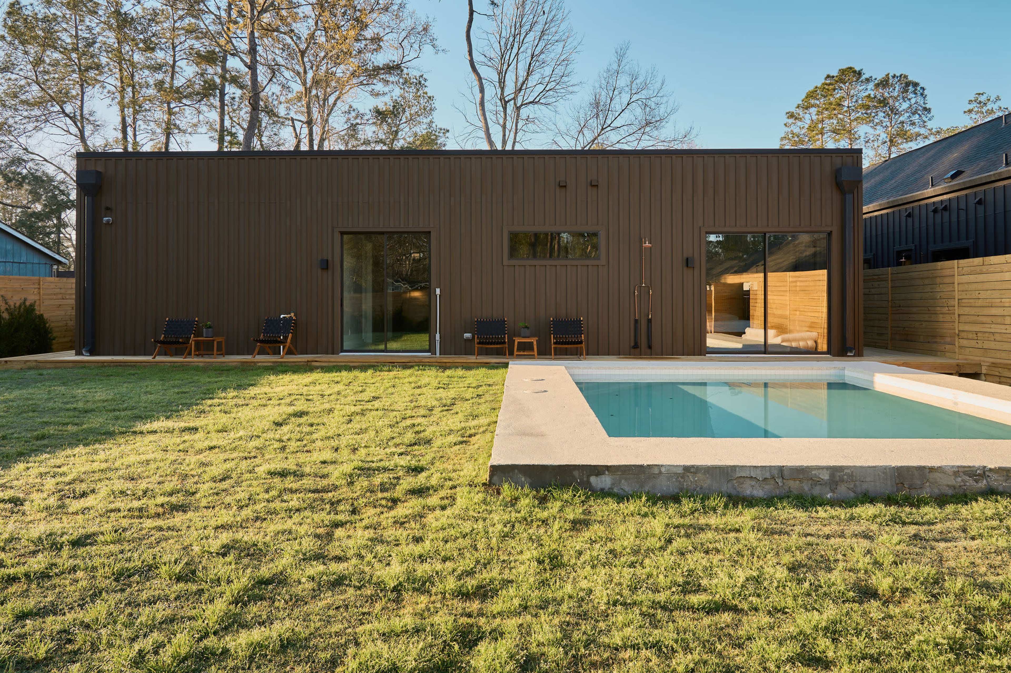The image shows a modern, minimalist house with a brown metal exterior, adjacent to a small rectangular swimming pool surrounded by a grassy lawn.