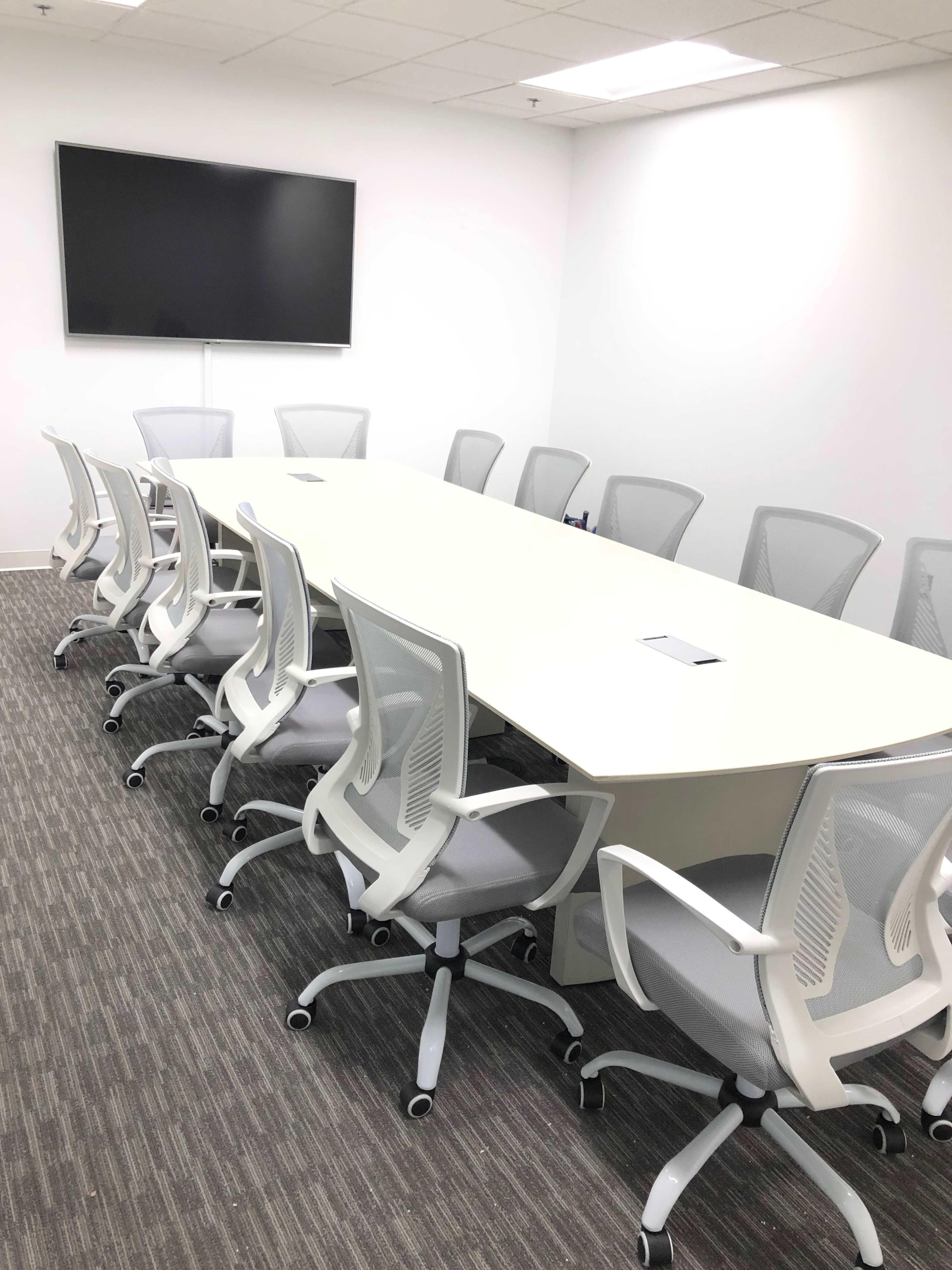 A modern conference room features a long white table surrounded by eight light gray chairs, with a wall-mounted television at one end.