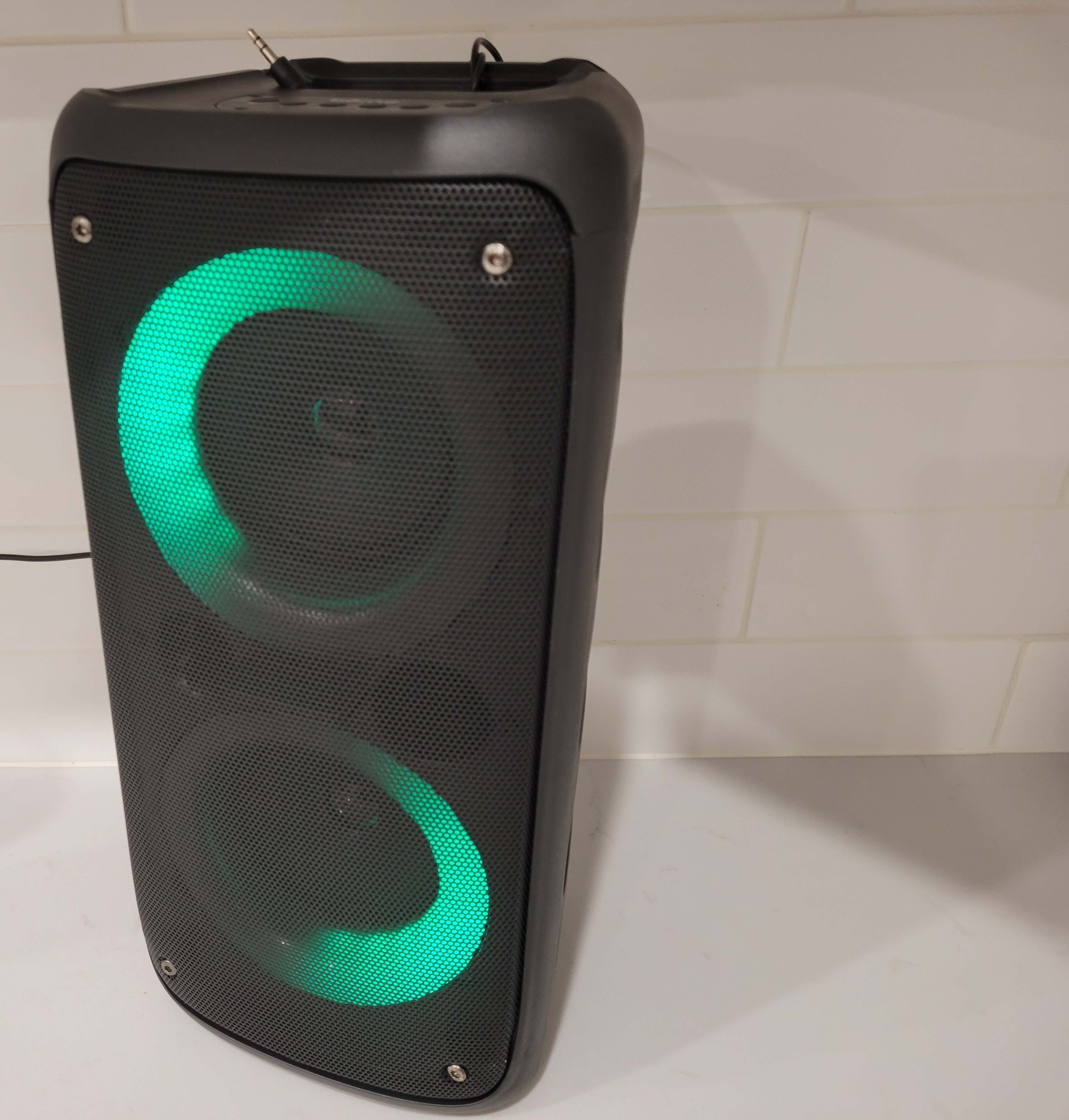 A black portable speaker with green lights around its two large speakers is placed on a white surface.