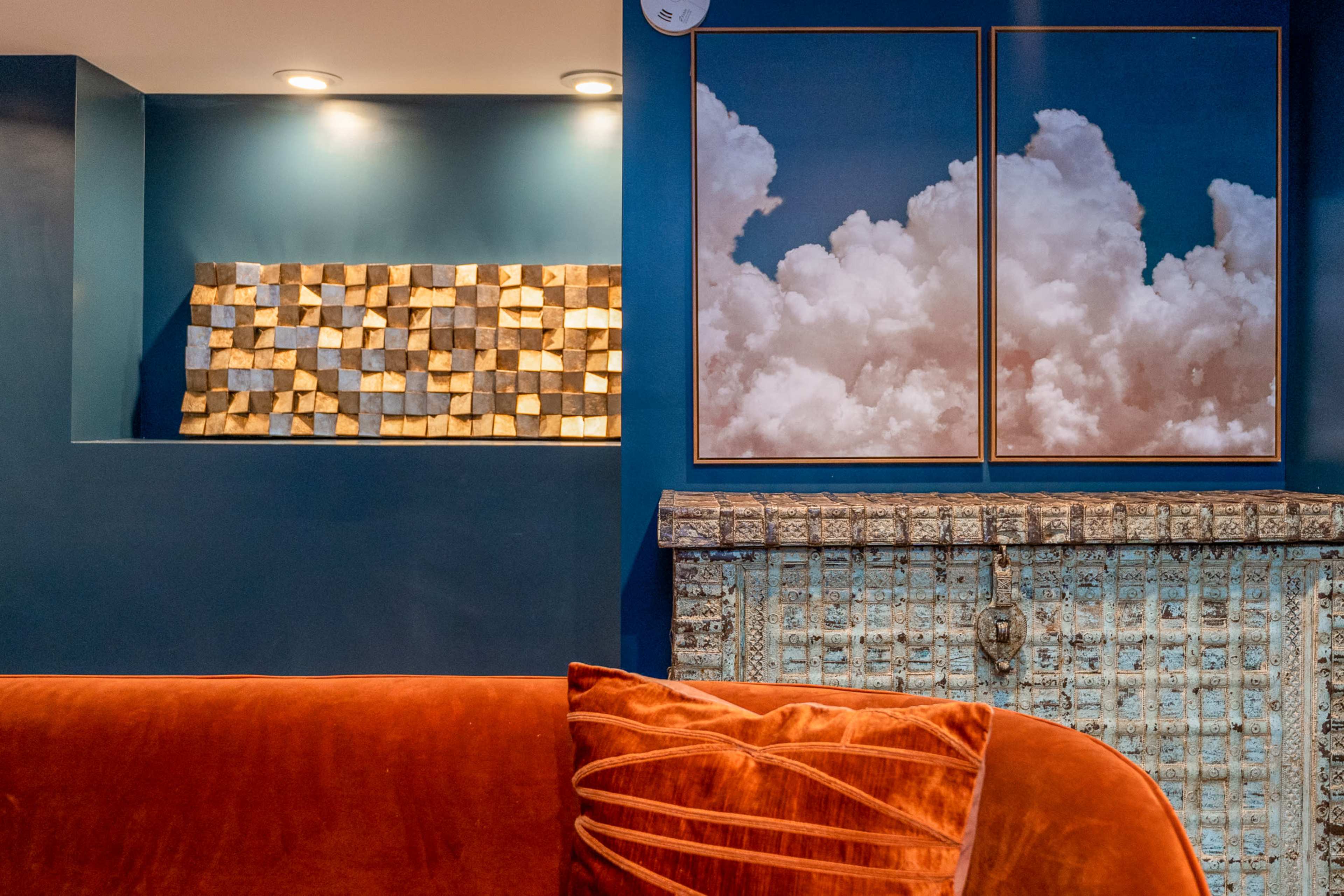 A living room with a deep blue wall featuring two framed cloud prints and a textured artwork made of cubes, complemented by an ornate mantel and an orange couch with decorative pillows.