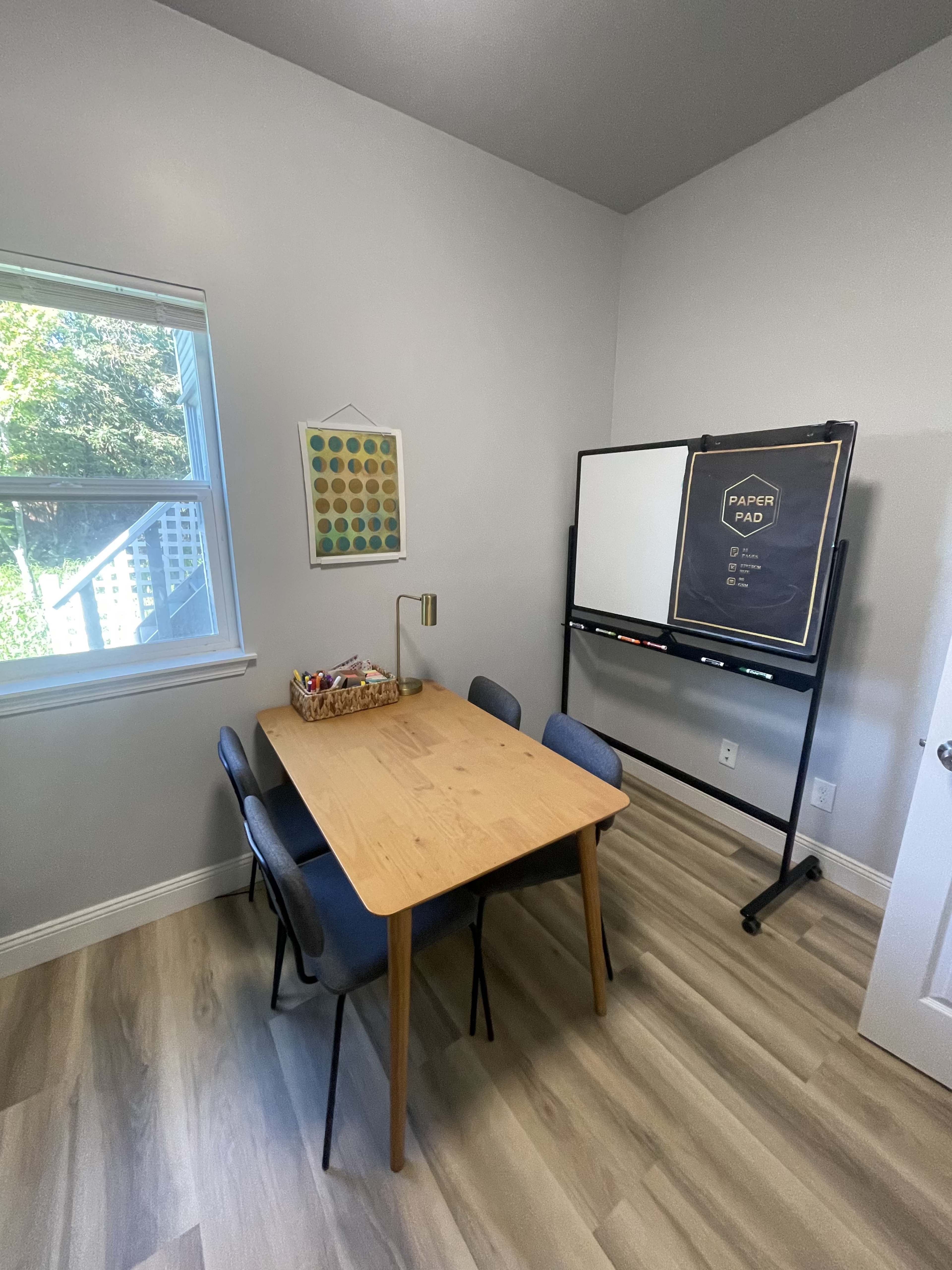 A small room with a wooden table and four chairs, a wall-mounted chart, and a whiteboard on a stand, with a window providing natural light.