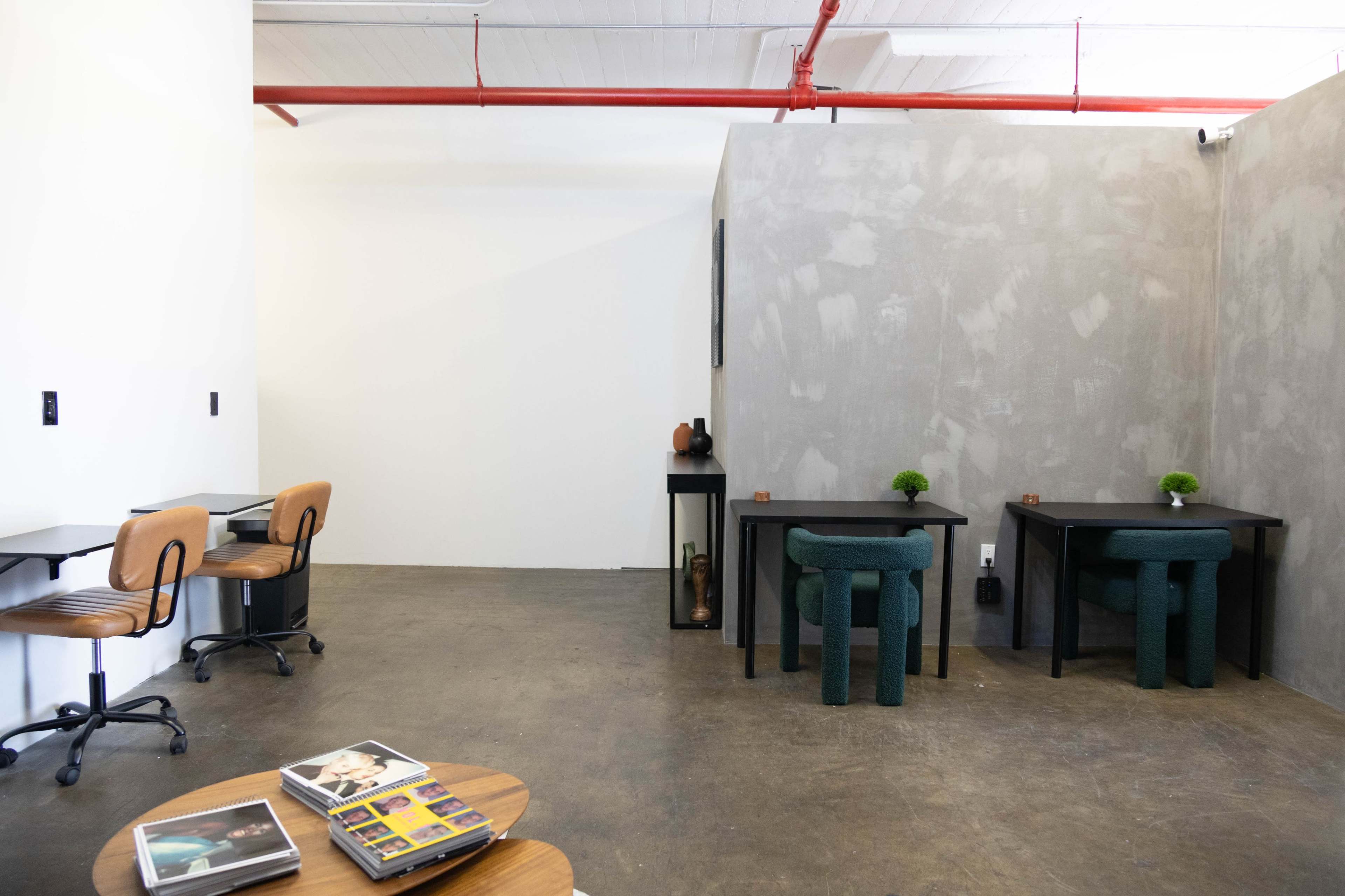 The image shows a minimalist interior space with two black tables, wooden chairs, and a gray wall, featuring a small plant and decorative items on the tables.