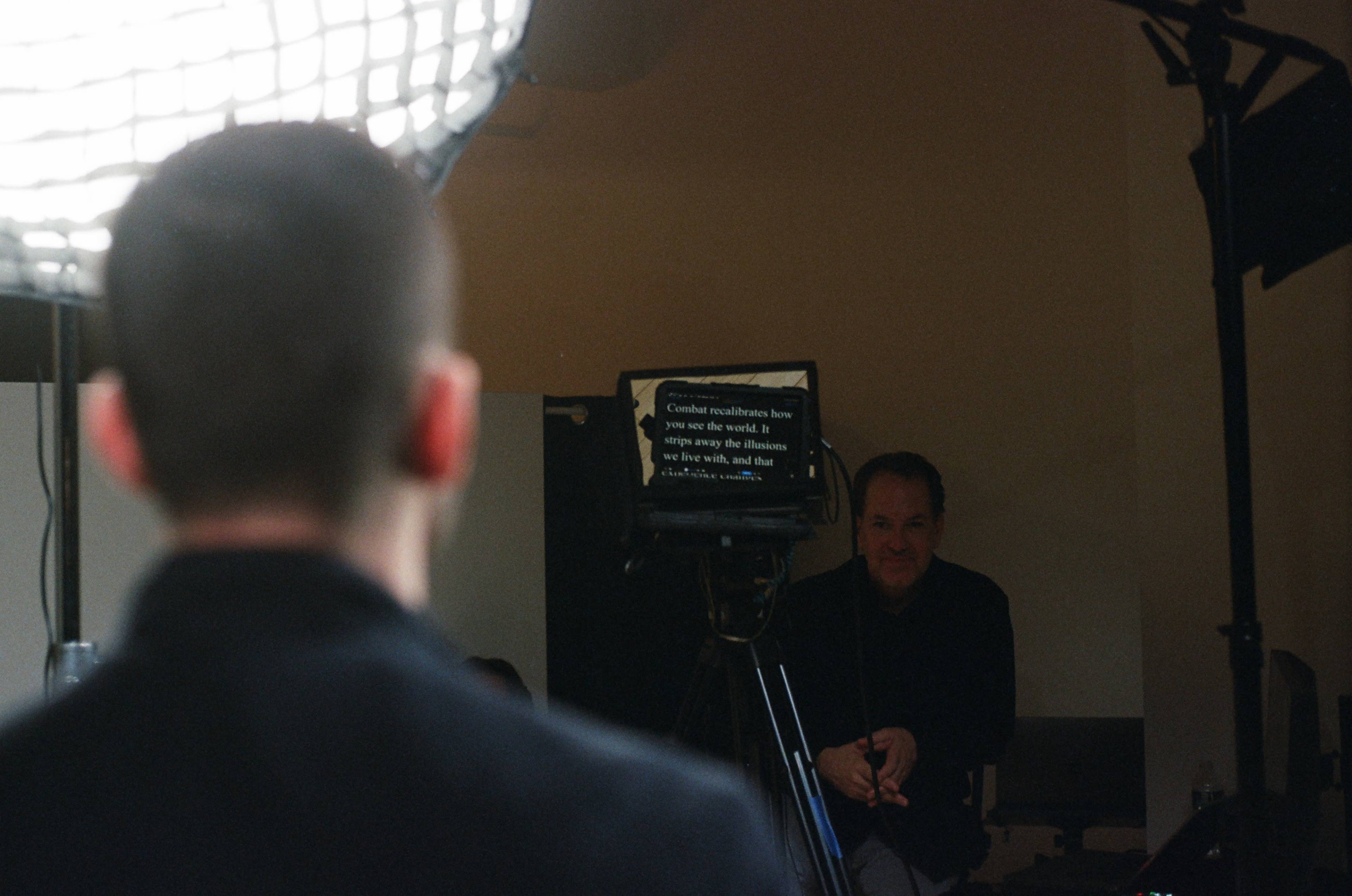 A person stands facing a camera setup while another person watches from behind a teleprompter.