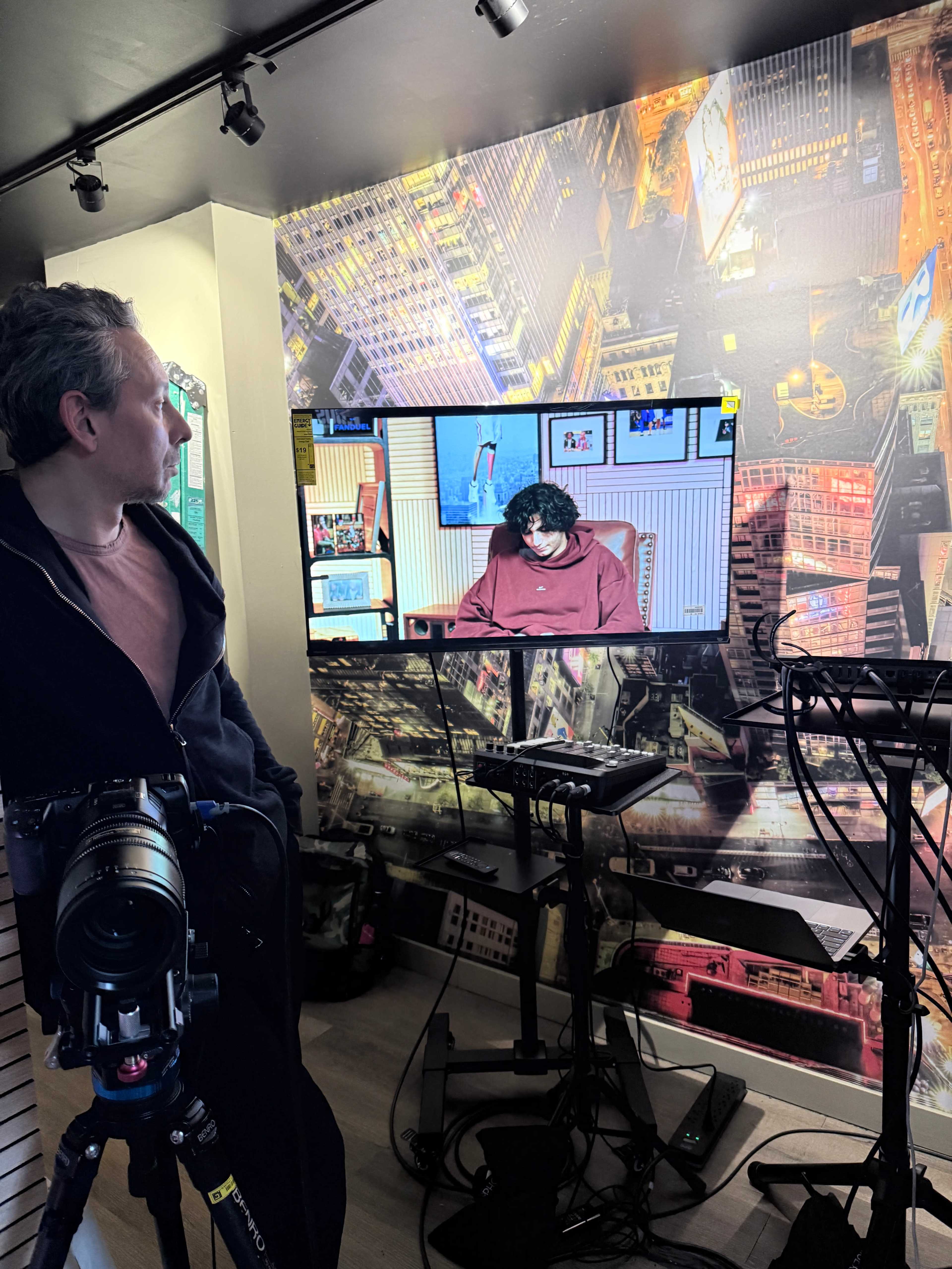 A man stands beside a seated person at a desk in a small studio, with cityscape wallpaper and a camera set up nearby.