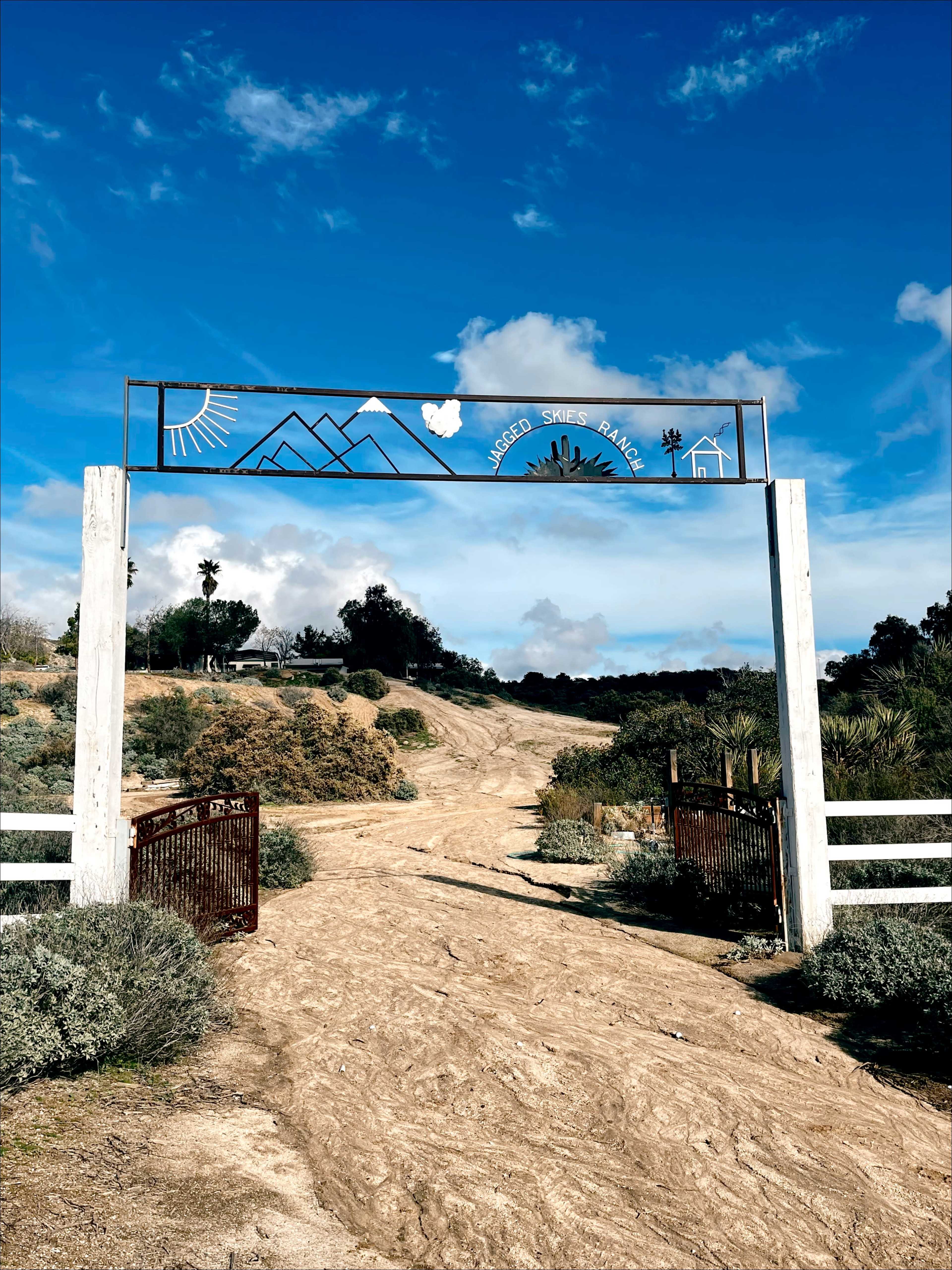 A weathered metal archway with a decorative design marking the entrance to a dirt path leading into a rural area surrounded by shrubs and trees under a blue sky.