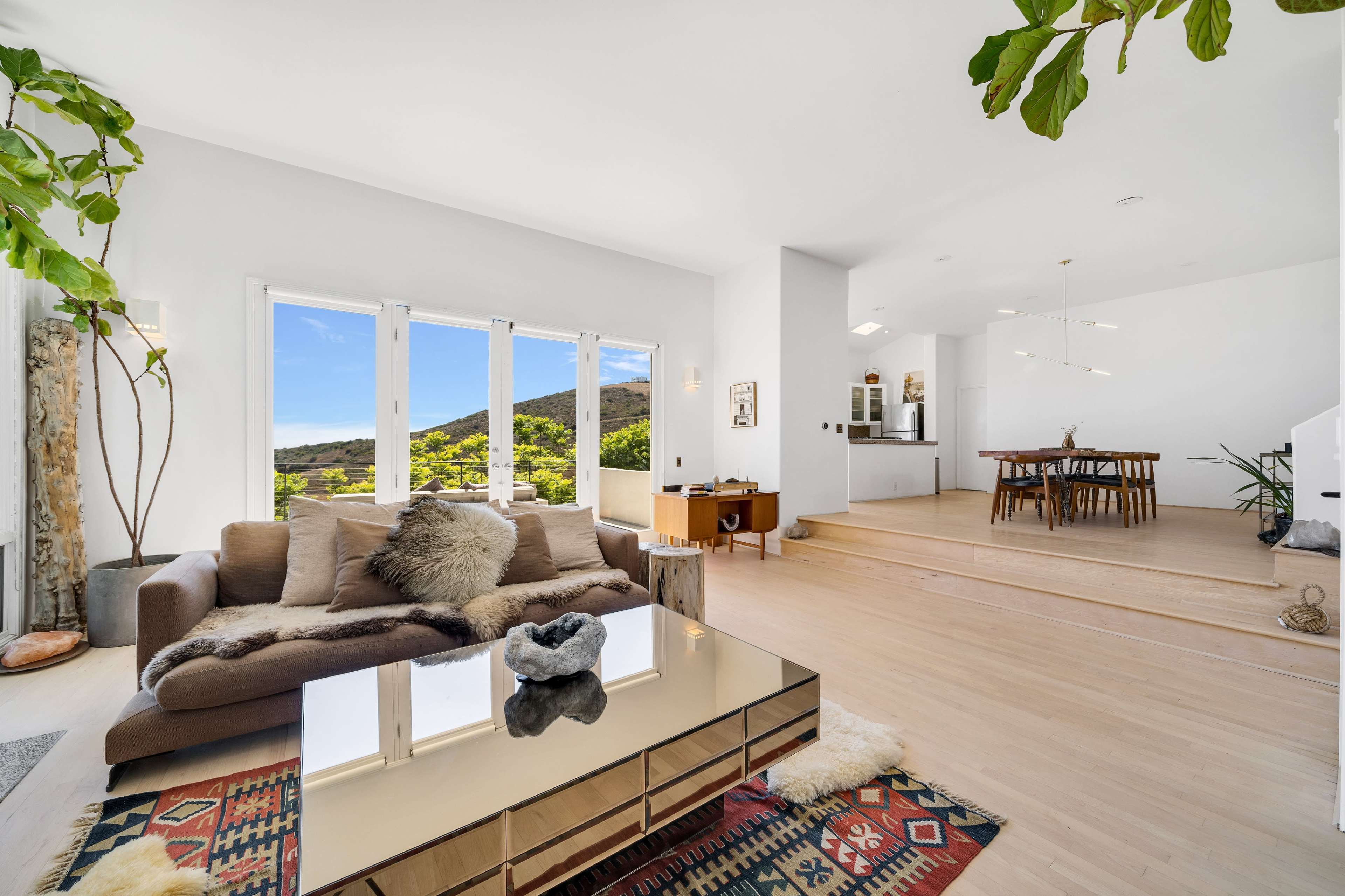 A bright, modern living room featuring a large window with a view of a green hillside, a sofa with decorative pillows, and a coffee table on a patterned rug.