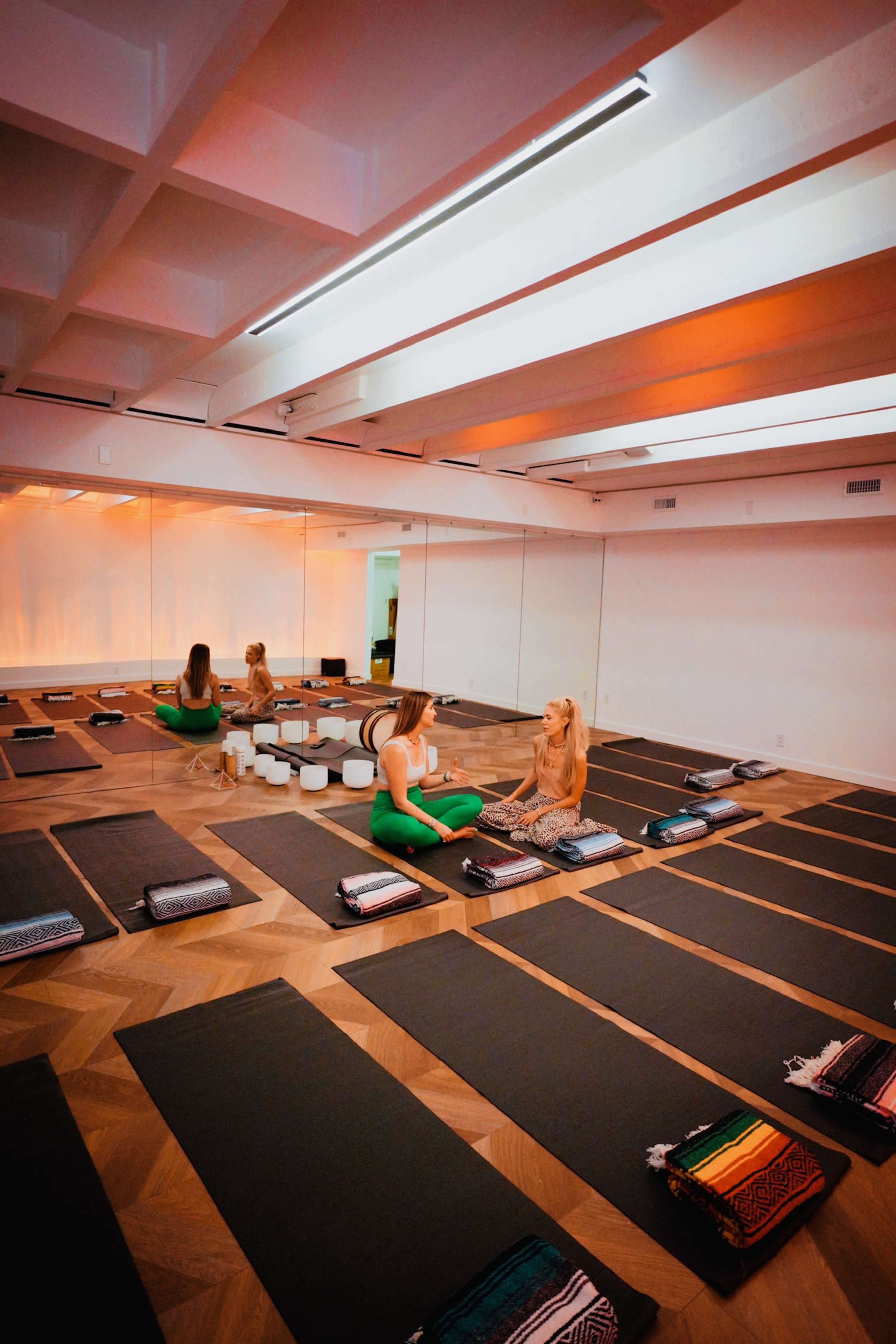 A yoga studio features several exercise mats arranged on a wooden floor, with two women sitting and conversing in a well-lit space.