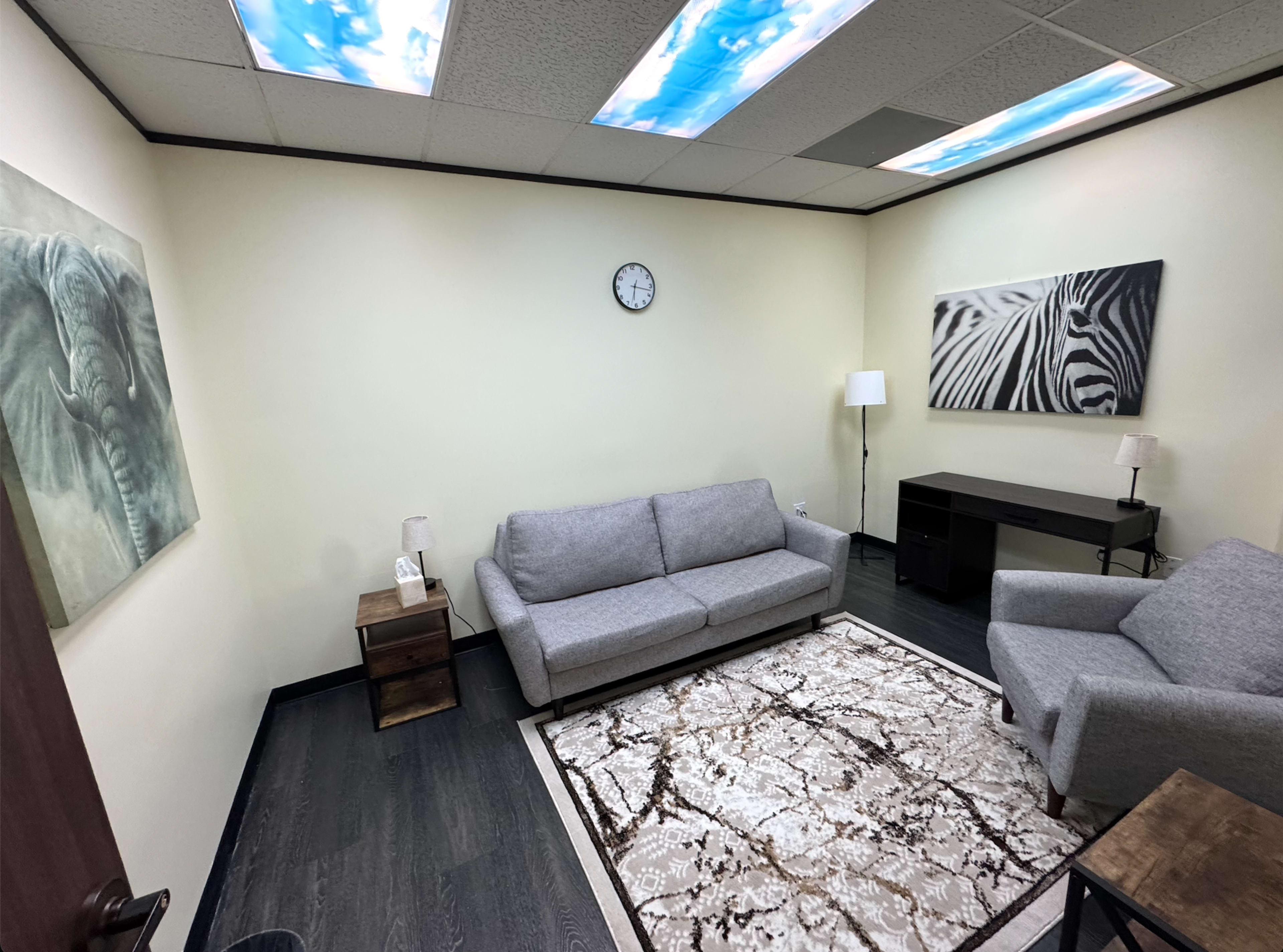 The room features two gray sofas, a coffee table, and animal-themed artwork on the walls, with a clock and lamps providing light.