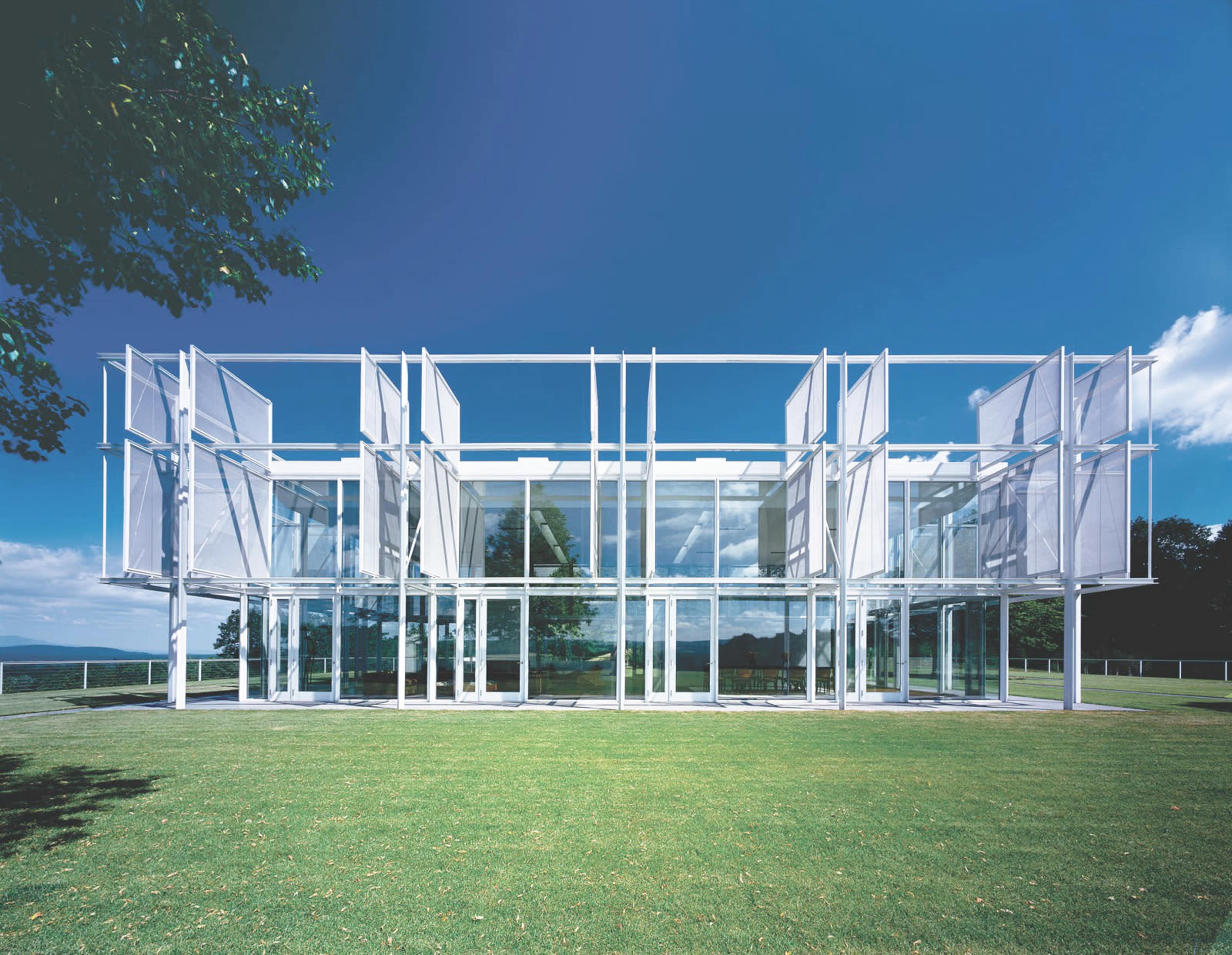 A modern, glass-fronted building with a white structural framework set on a grassy area under a clear blue sky.