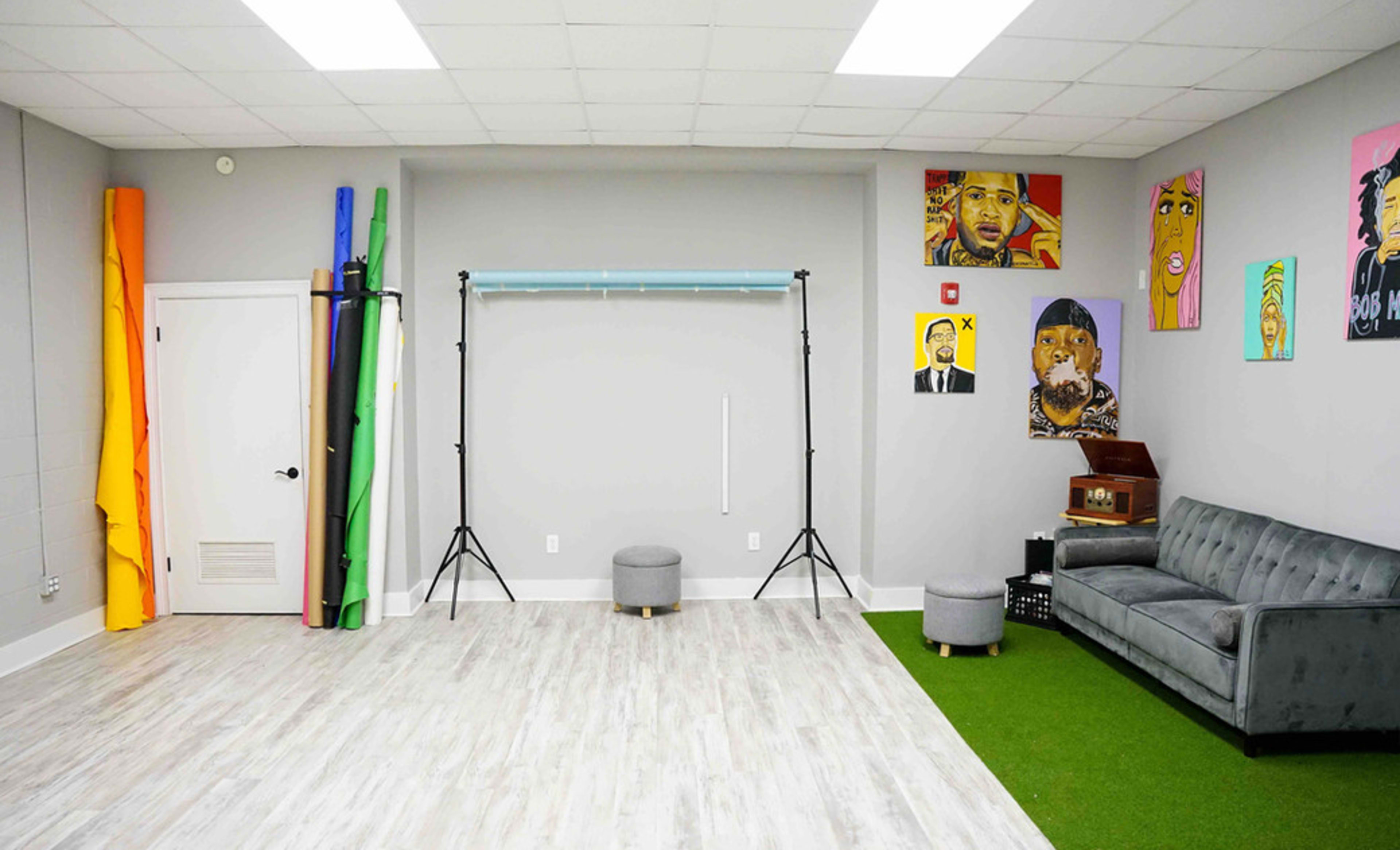 A spacious studio with colorful backdrop rolls, a photography setup, artwork on the walls, a vinyl record player, and a gray couch on a green area rug.