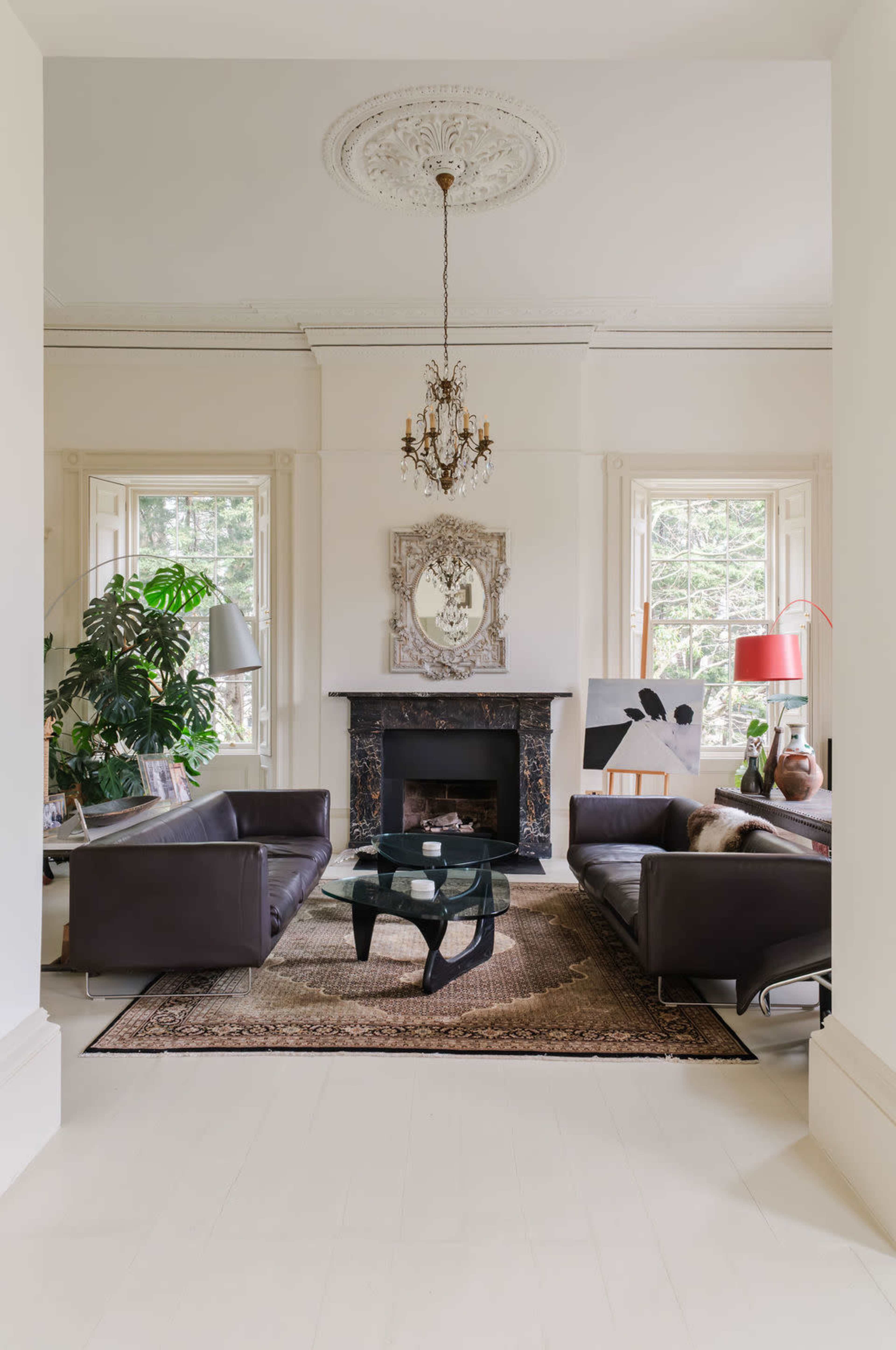 The image shows a spacious living room with two dark leather sofas facing a glass coffee table, a decorative fireplace, and large windows that allow natural light to enter the space.