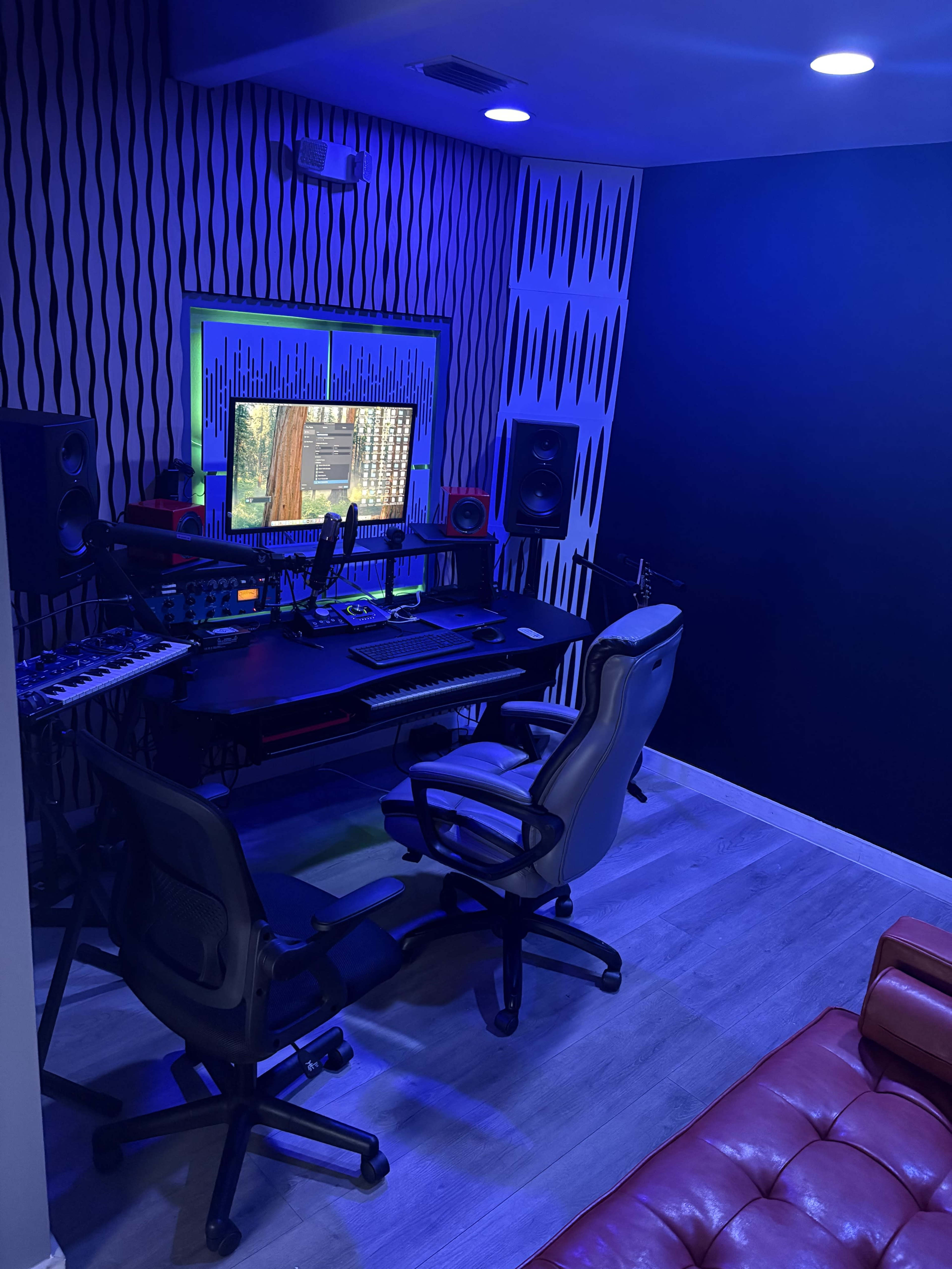 The image shows a music studio featuring a black workstation with audio equipment, two office chairs, and blue lighting on the walls.