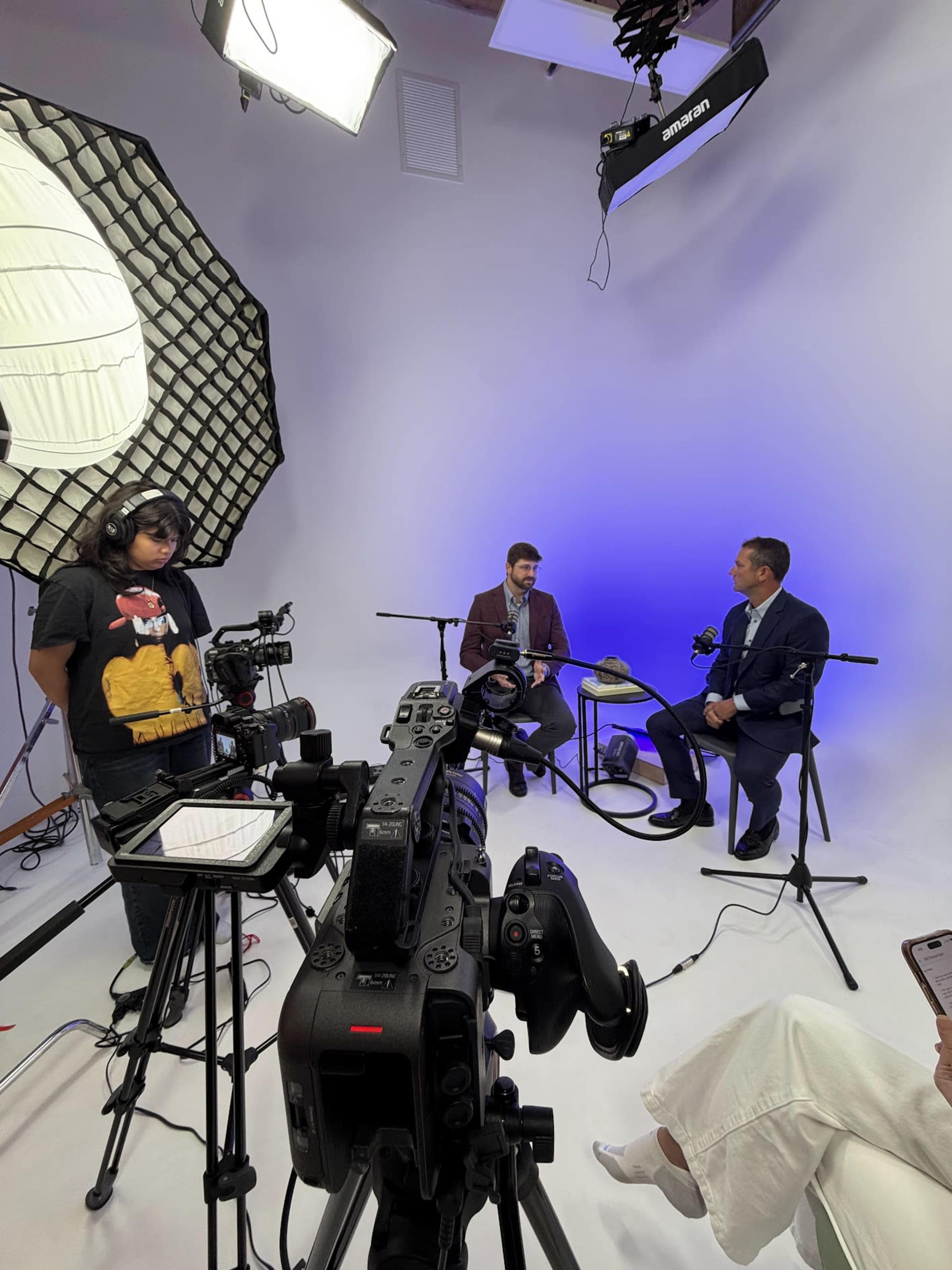 Two men are seated in a studio setting with multiple cameras and lighting equipment, while a crew member adjusts the lighting.