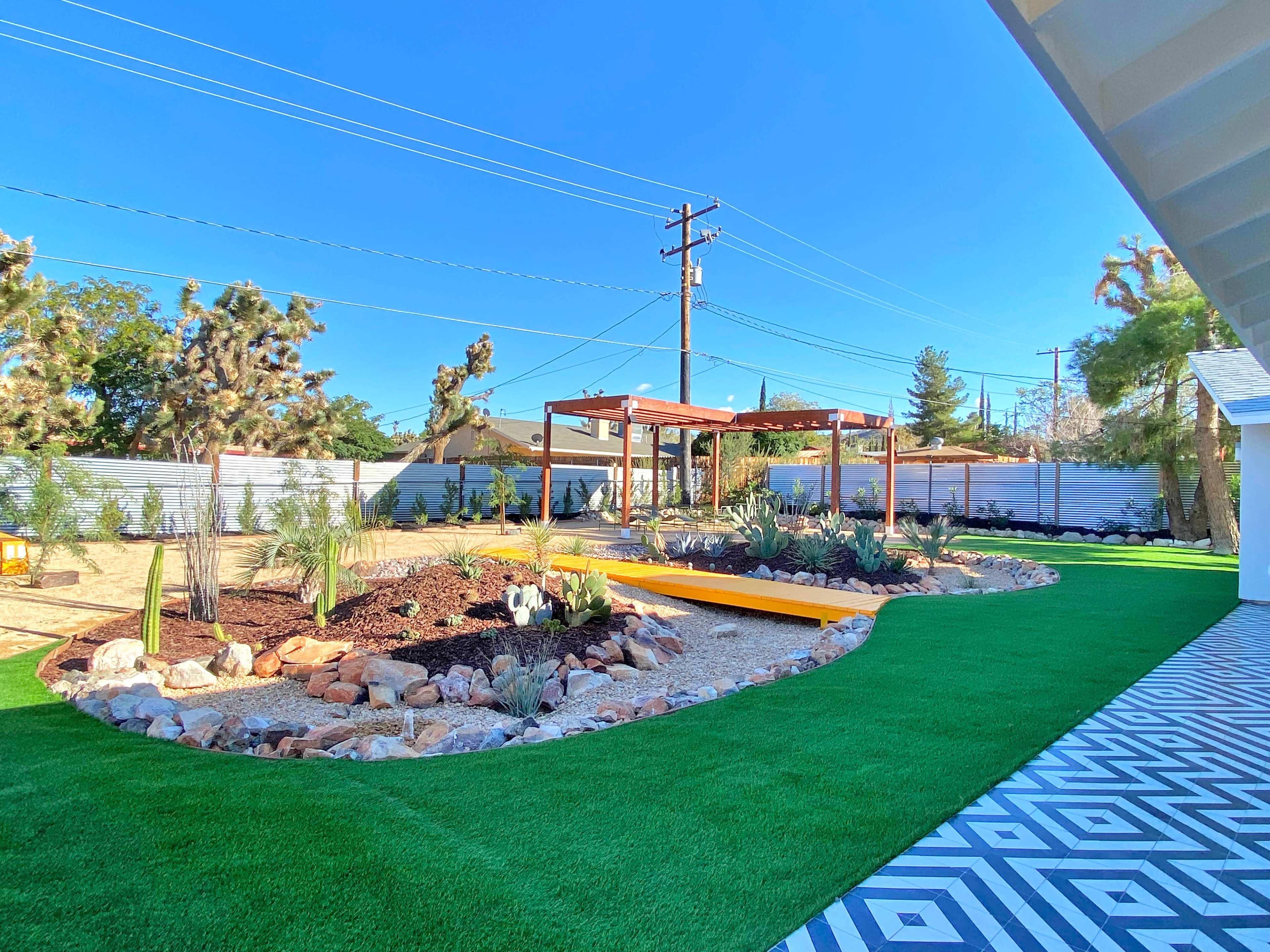 The image shows a landscaped backyard featuring a grassy area, a rock garden, wooden pergolas, and a pathway with patterned tiles.