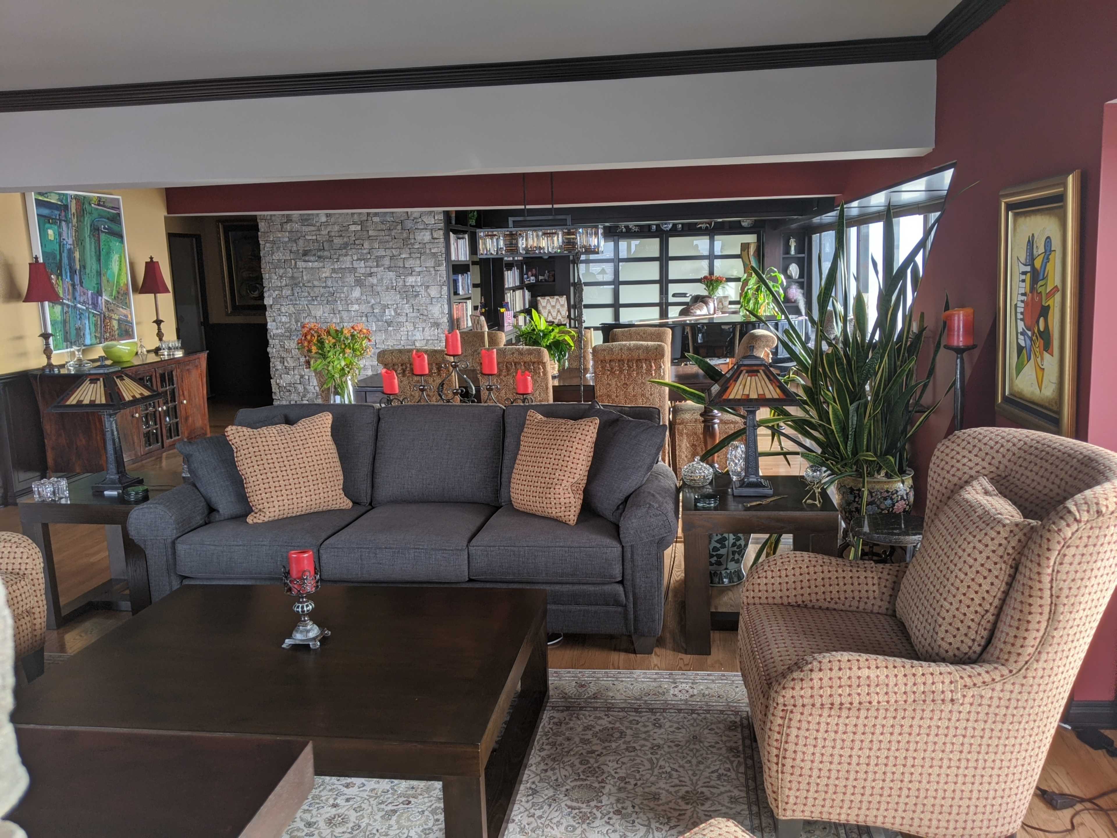 A modern living room with a gray sofa, patterned armchairs, a wooden coffee table, and plants, all set against a backdrop of large windows and a stone wall.