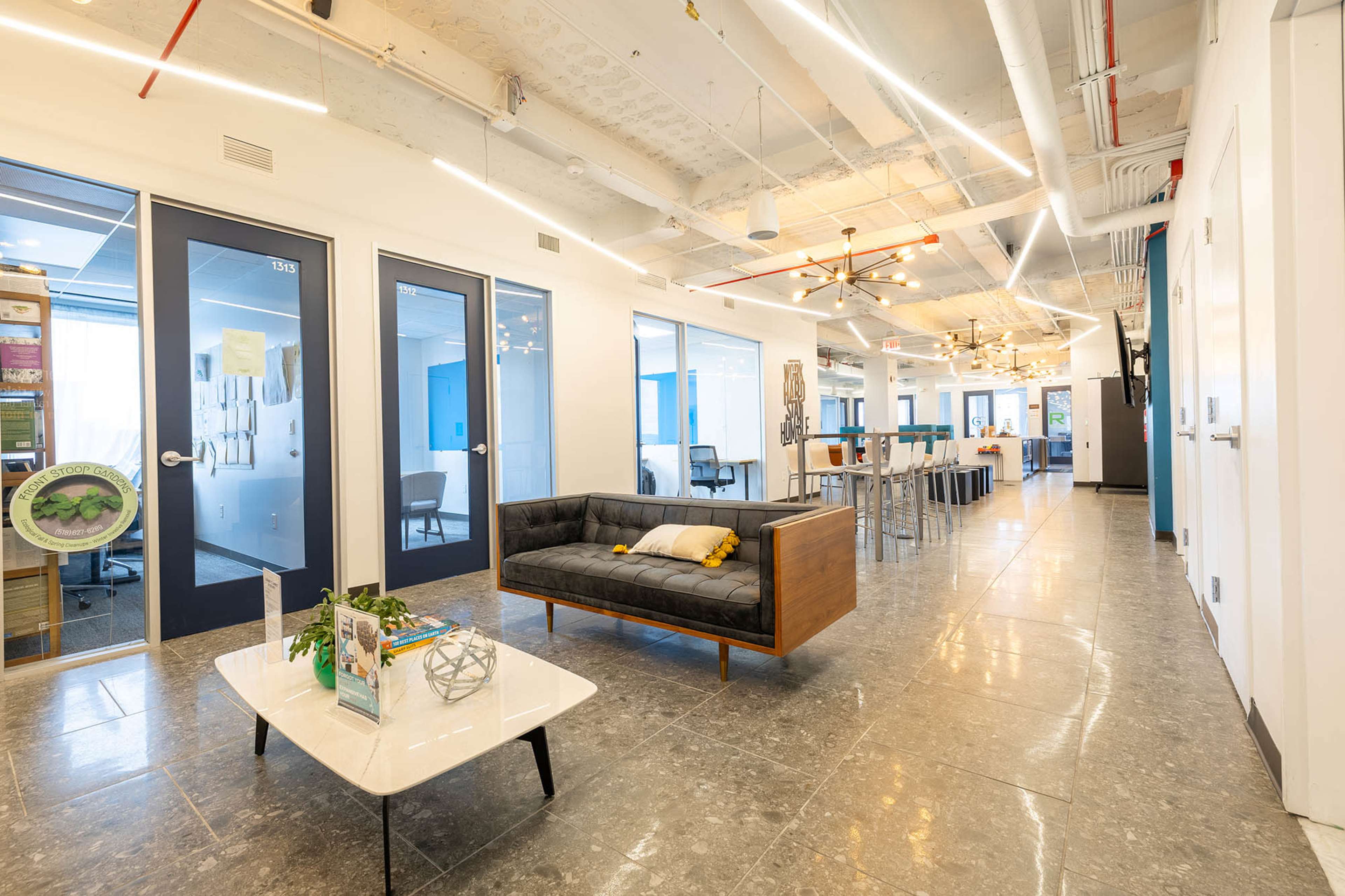 The image shows a modern office interior with a seating area, glass-walled meeting rooms, and a collaborative workspace featuring tall tables and chairs.