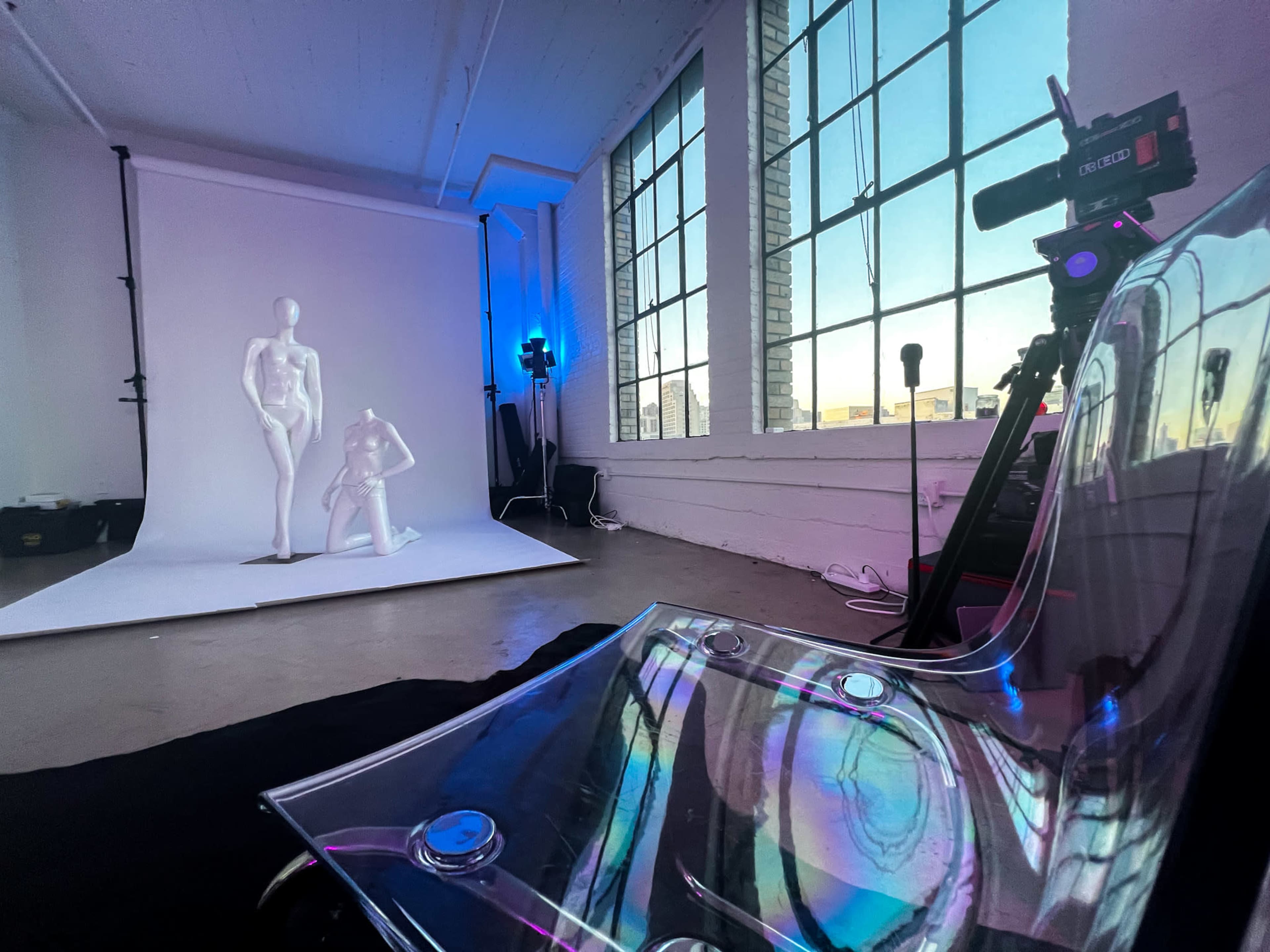 A clear chair sits in the foreground of a photography studio featuring a backdrop with two mannequins and equipment for a photoshoot.