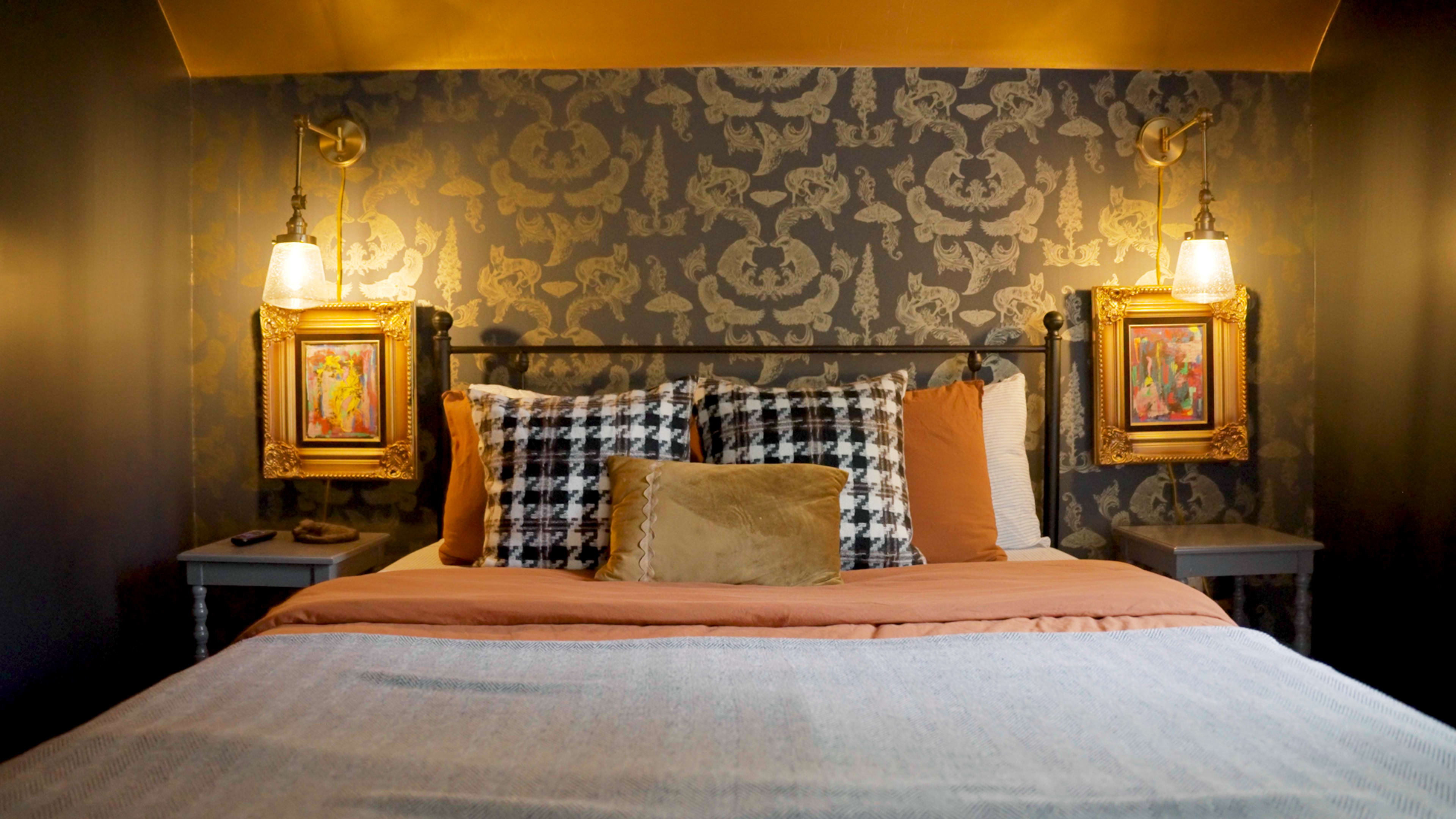 The image shows a cozy bedroom featuring a bed with patterned pillows and blankets, framed artwork on the walls, and decorative wall sconces.