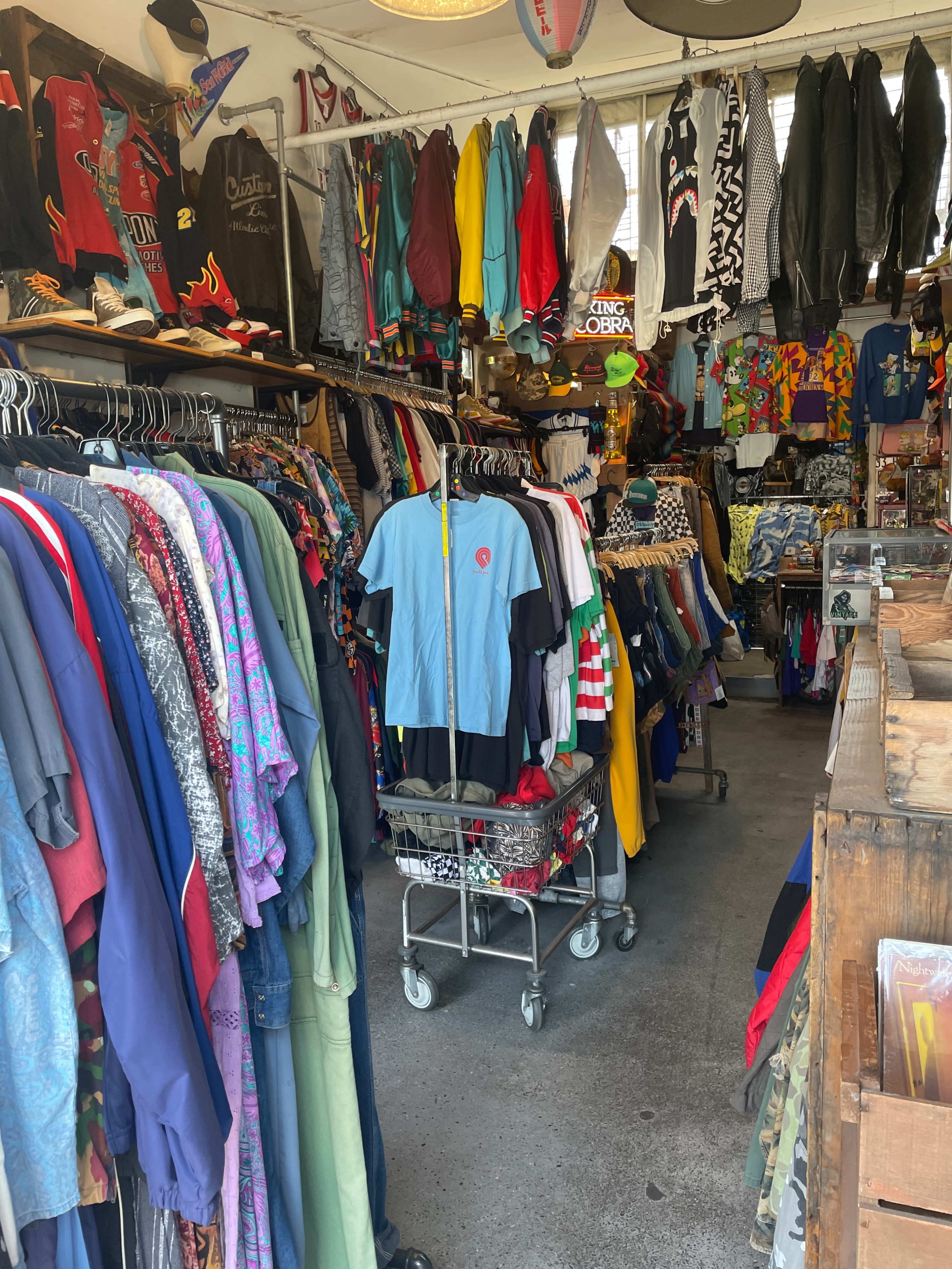 The image shows a clothing store densely packed with various garments hanging on racks, alongside a shopping cart filled with items.