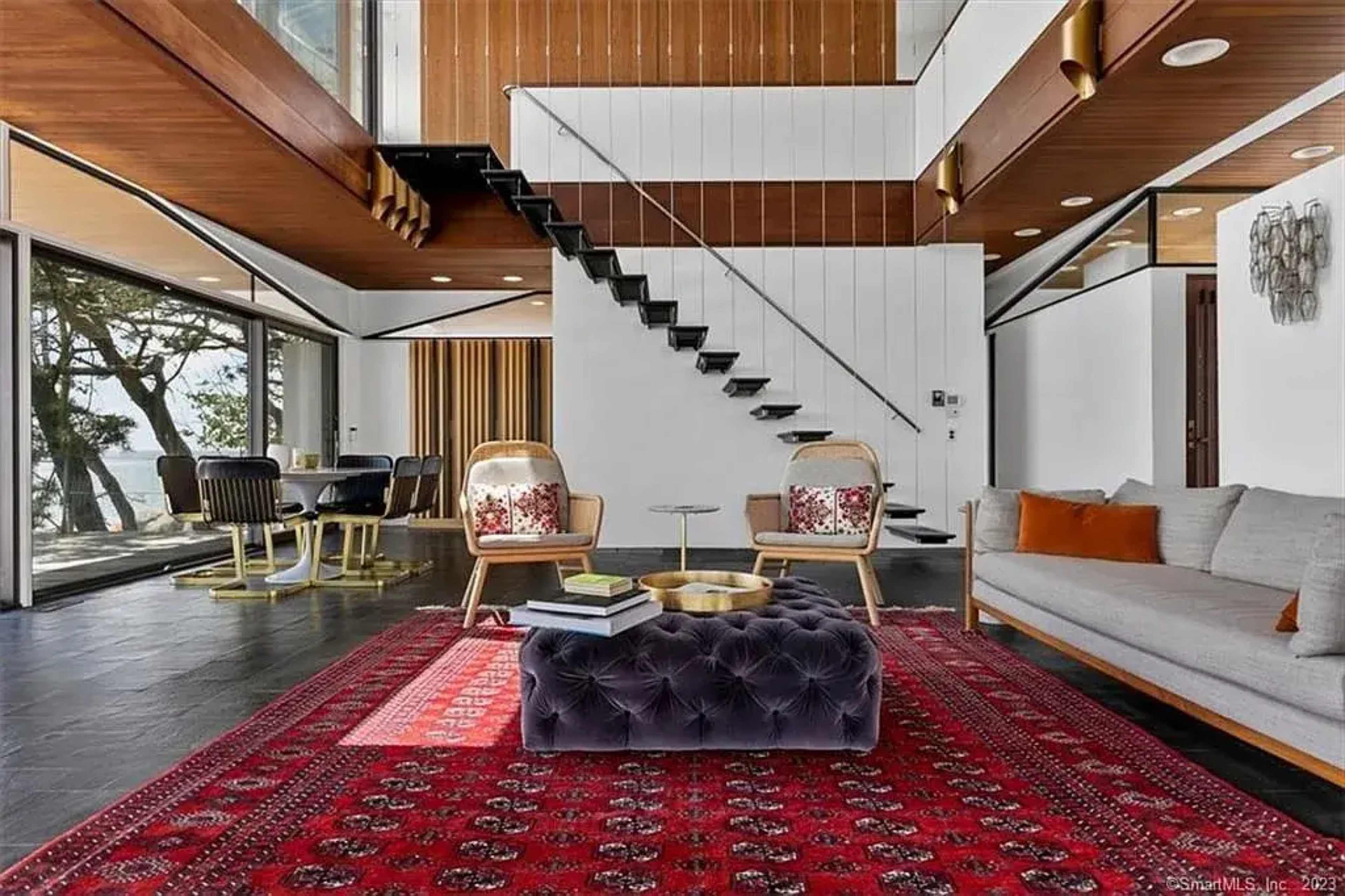 The image shows a modern living area featuring a gray sofa, a large red patterned rug, a glass wall with a view of trees, and a staircase leading to an upper level.