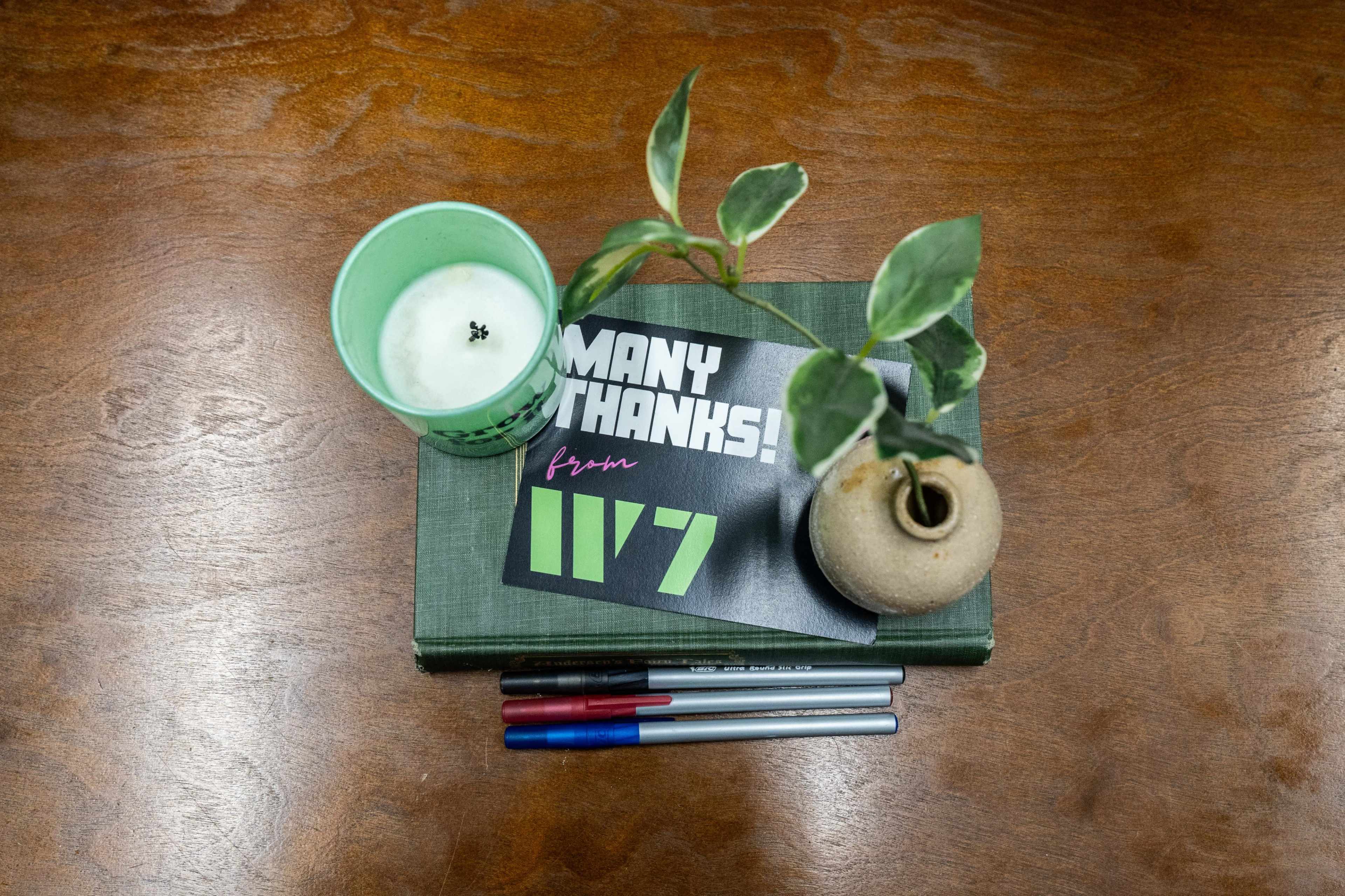 A green cup with a candle, a "THANKS" note, a small vase with a plant, and pens are arranged on a wooden table.
