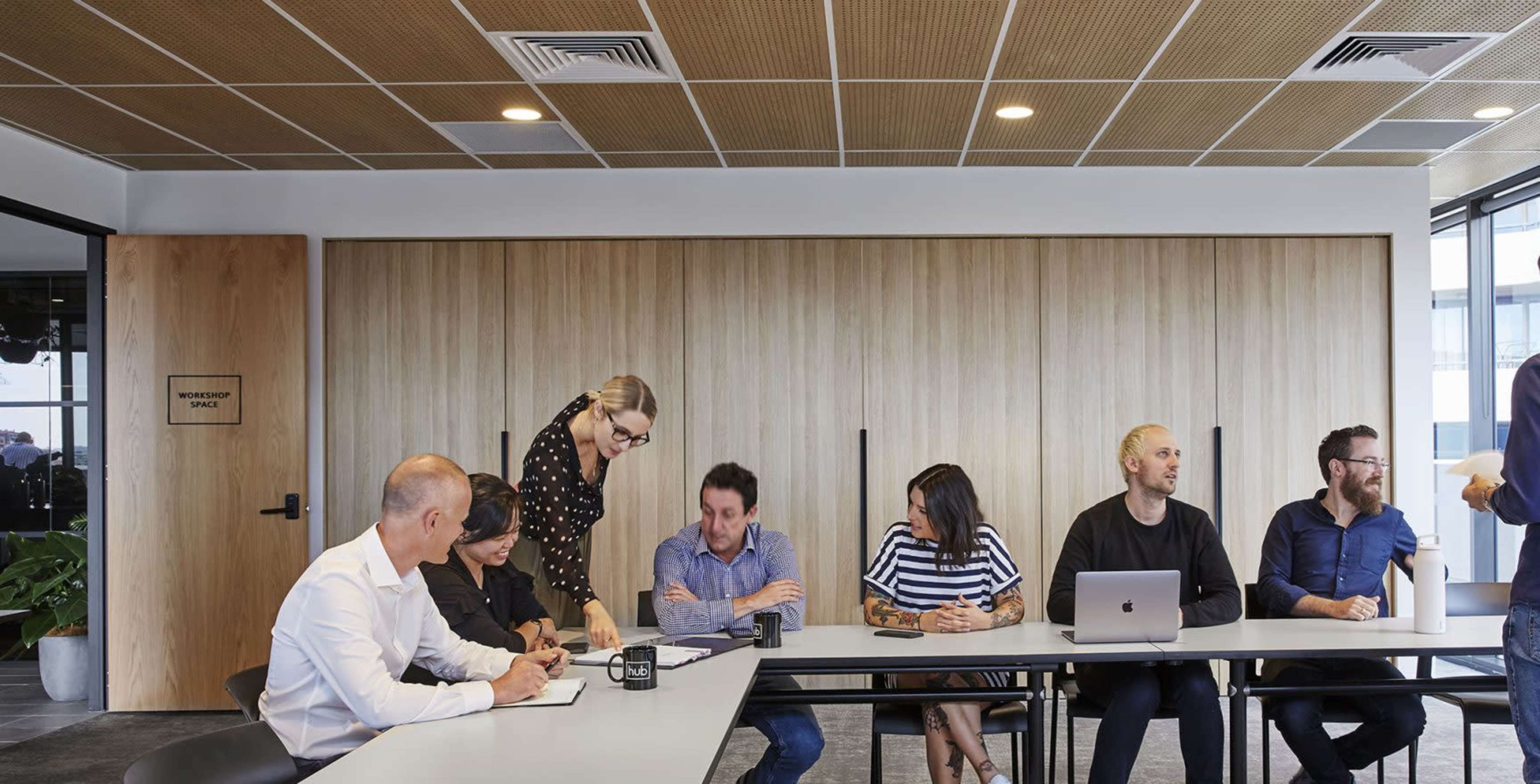 A group of professionals is seated around a long table in a meeting room, while one person stands and addresses the group.