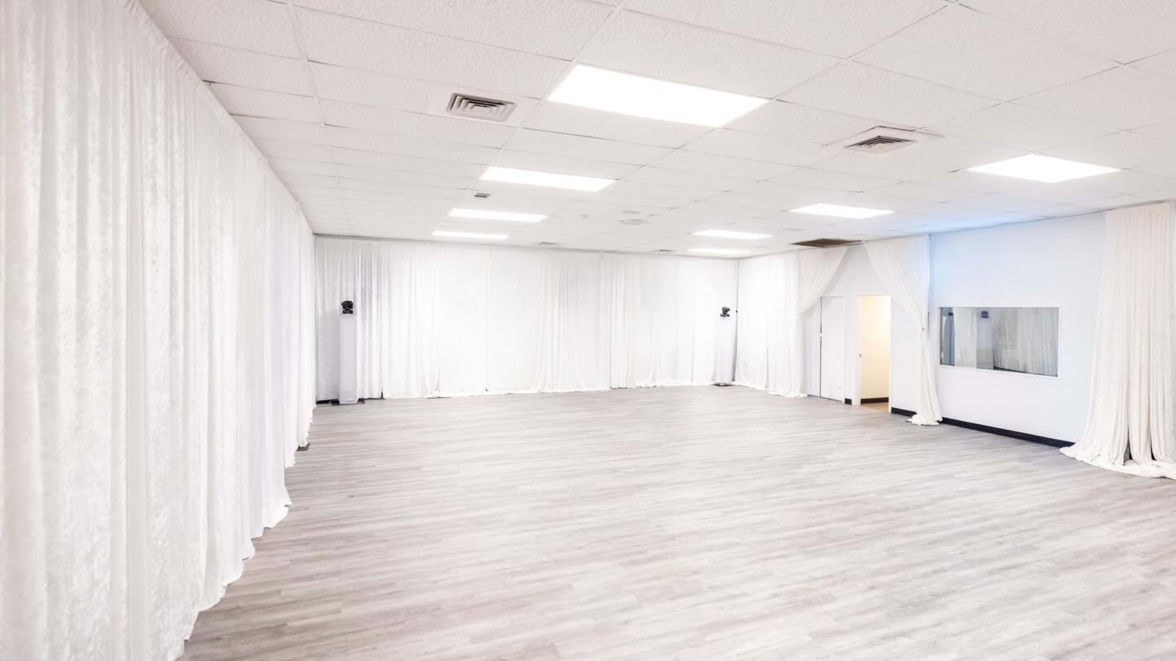 The image shows a spacious, empty room with white draped curtains along the walls and a light-colored wooden floor.