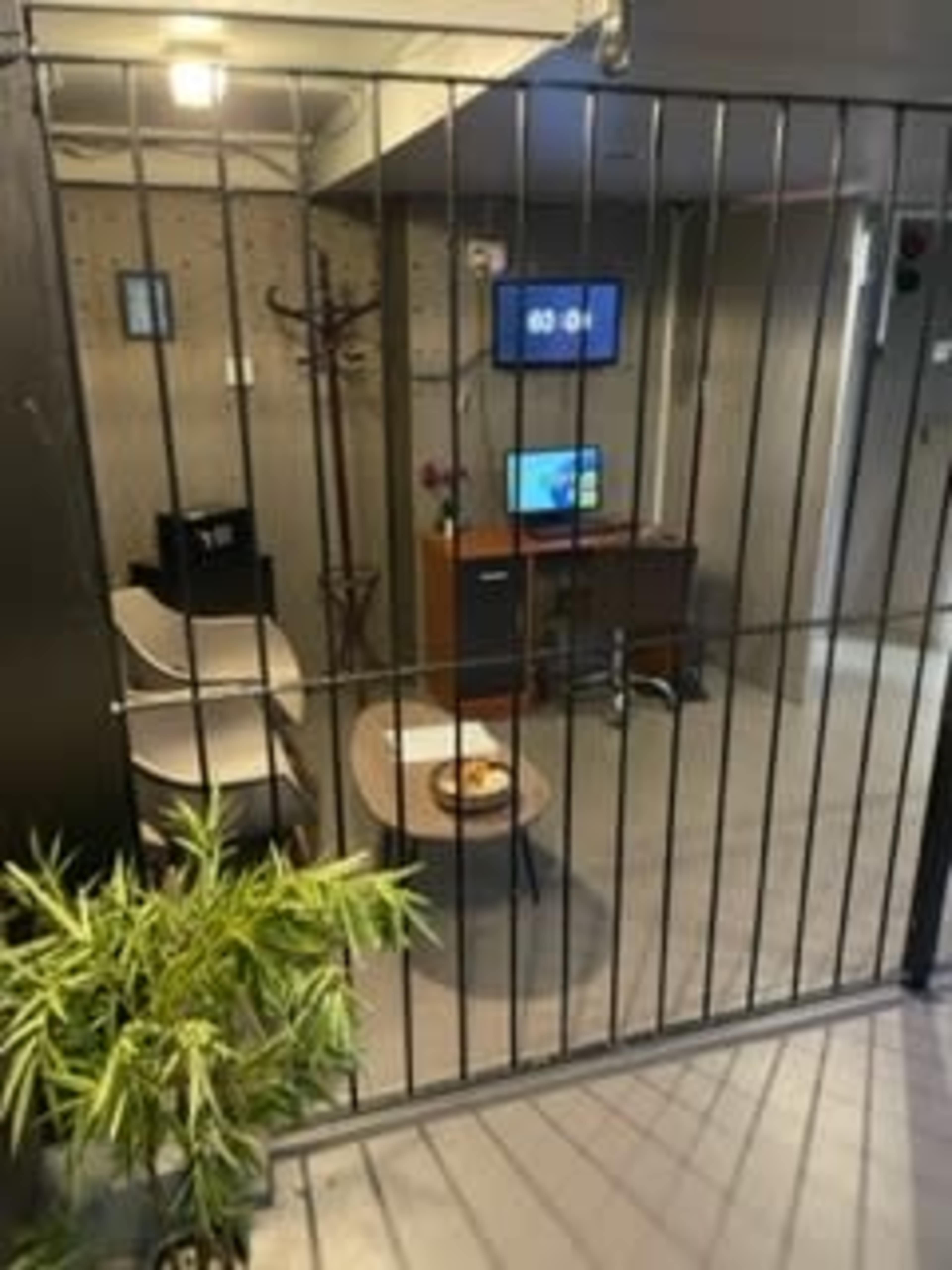 A small, enclosed office space with a desk, two chairs, a television, and a potted plant, all behind a metal grid barrier.