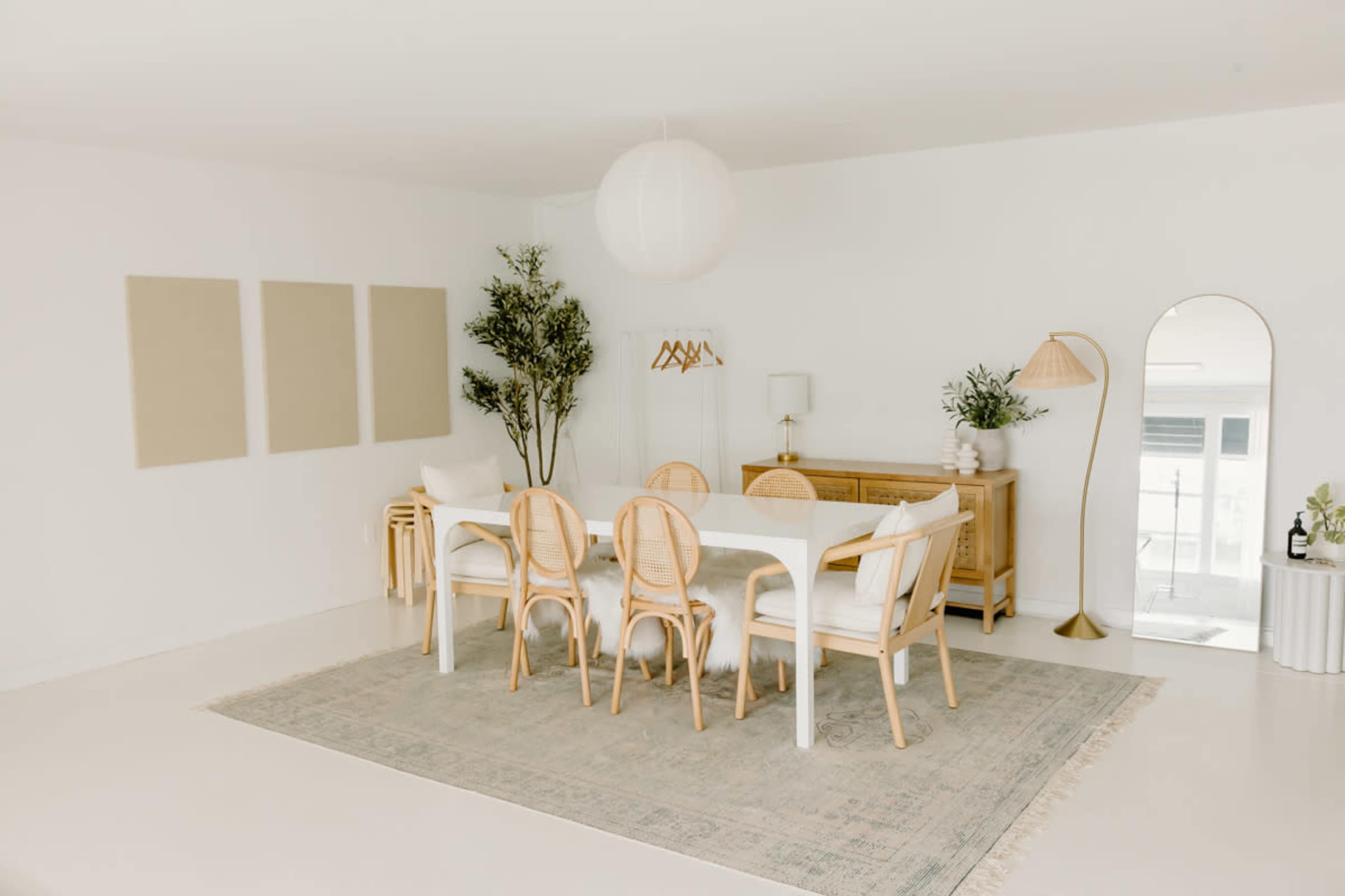 The image shows a bright, minimalist dining area featuring a white table surrounded by six chairs, a patterned rug, and a tall mirror, with decor including plants and wall hangings.