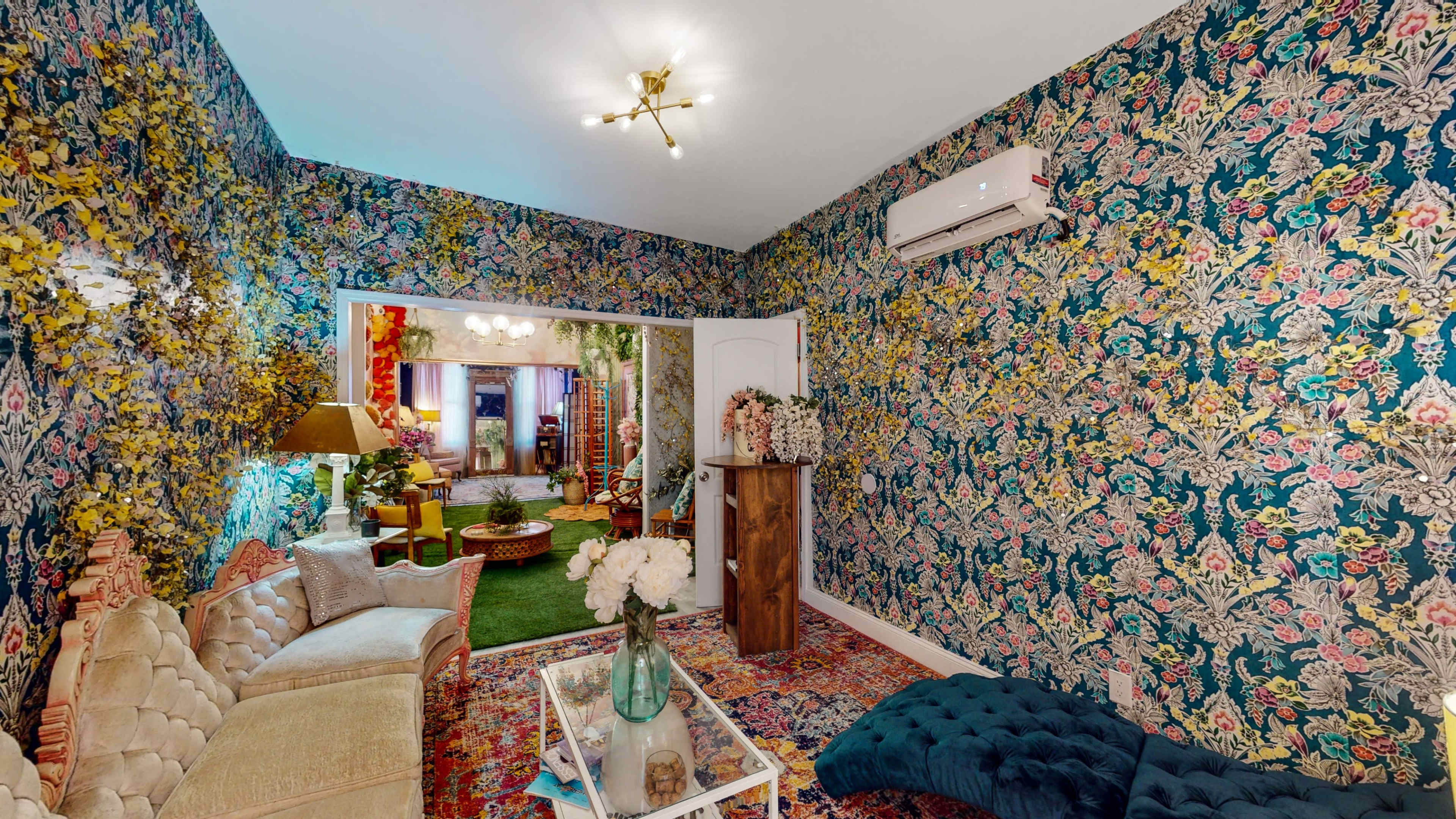 The room features floral wallpaper, a vintage sofa, a glass coffee table with decorative items, and a view of a garden through an open doorway.
