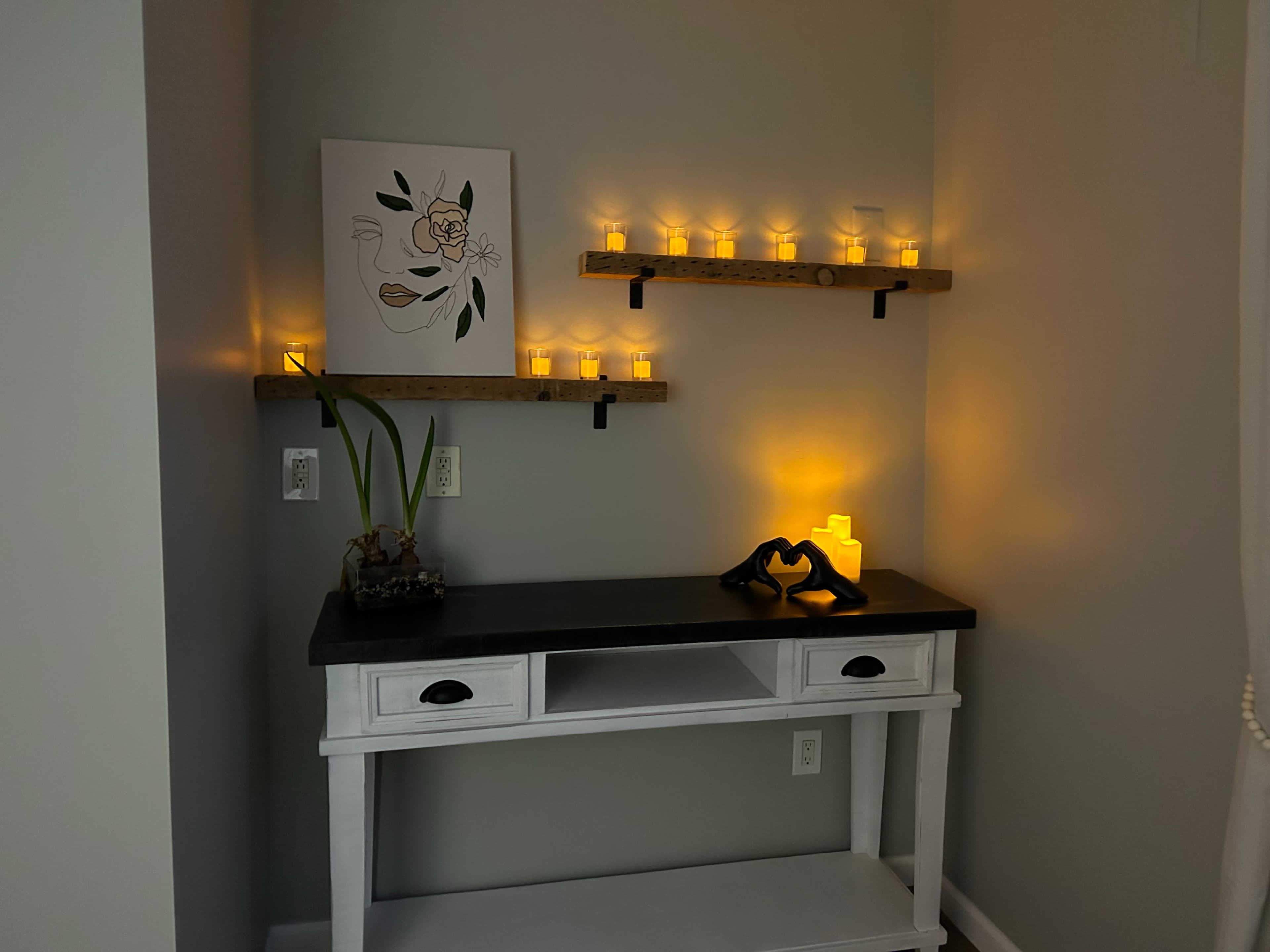 A white console table with a black top is positioned against a light grey wall, decorated with a minimalist art piece and illuminated candles on wooden shelves above.