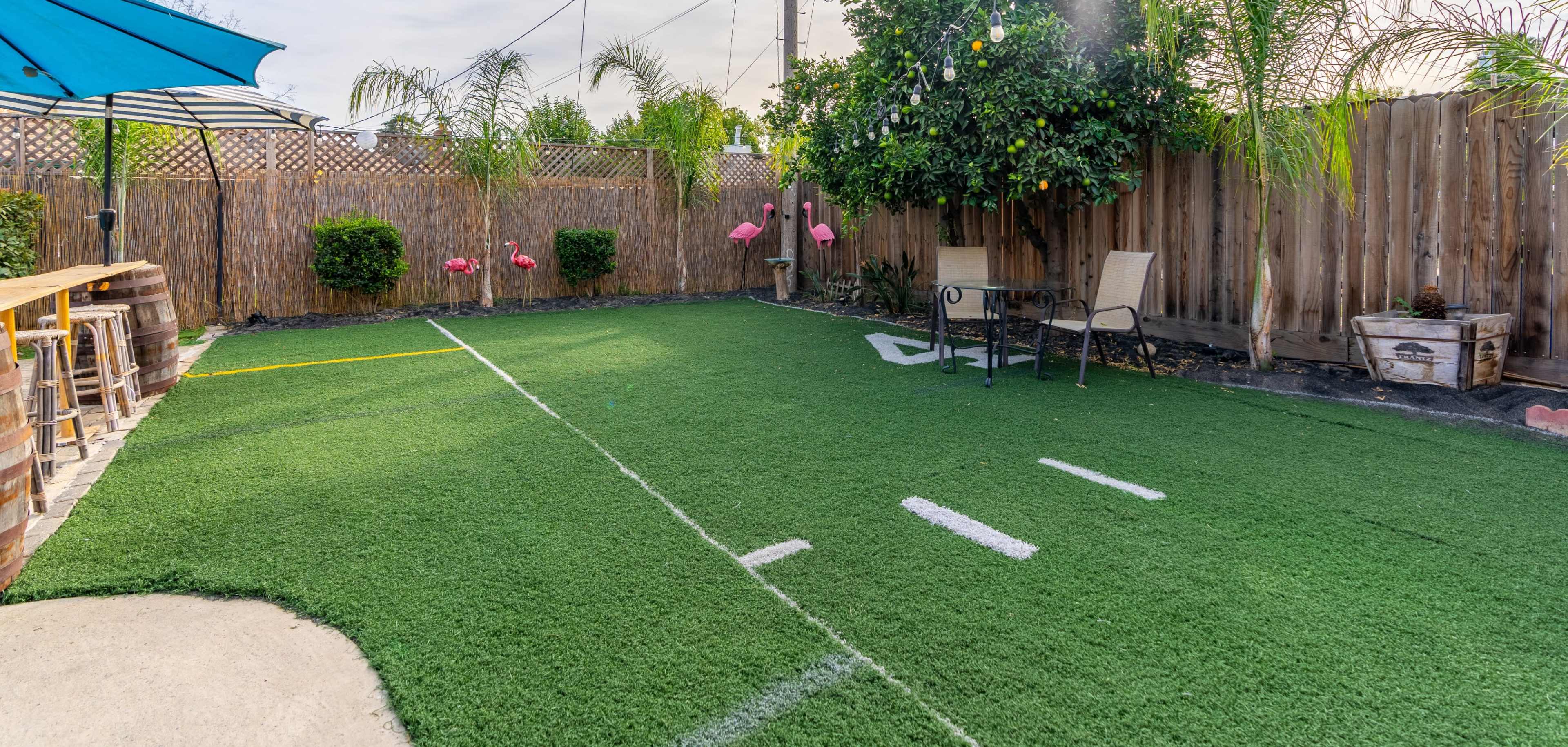 A backyard features a green artificial turf area designed like a football field, with pink flamingo decorations, a seating arrangement, and a bar counter under an umbrella.