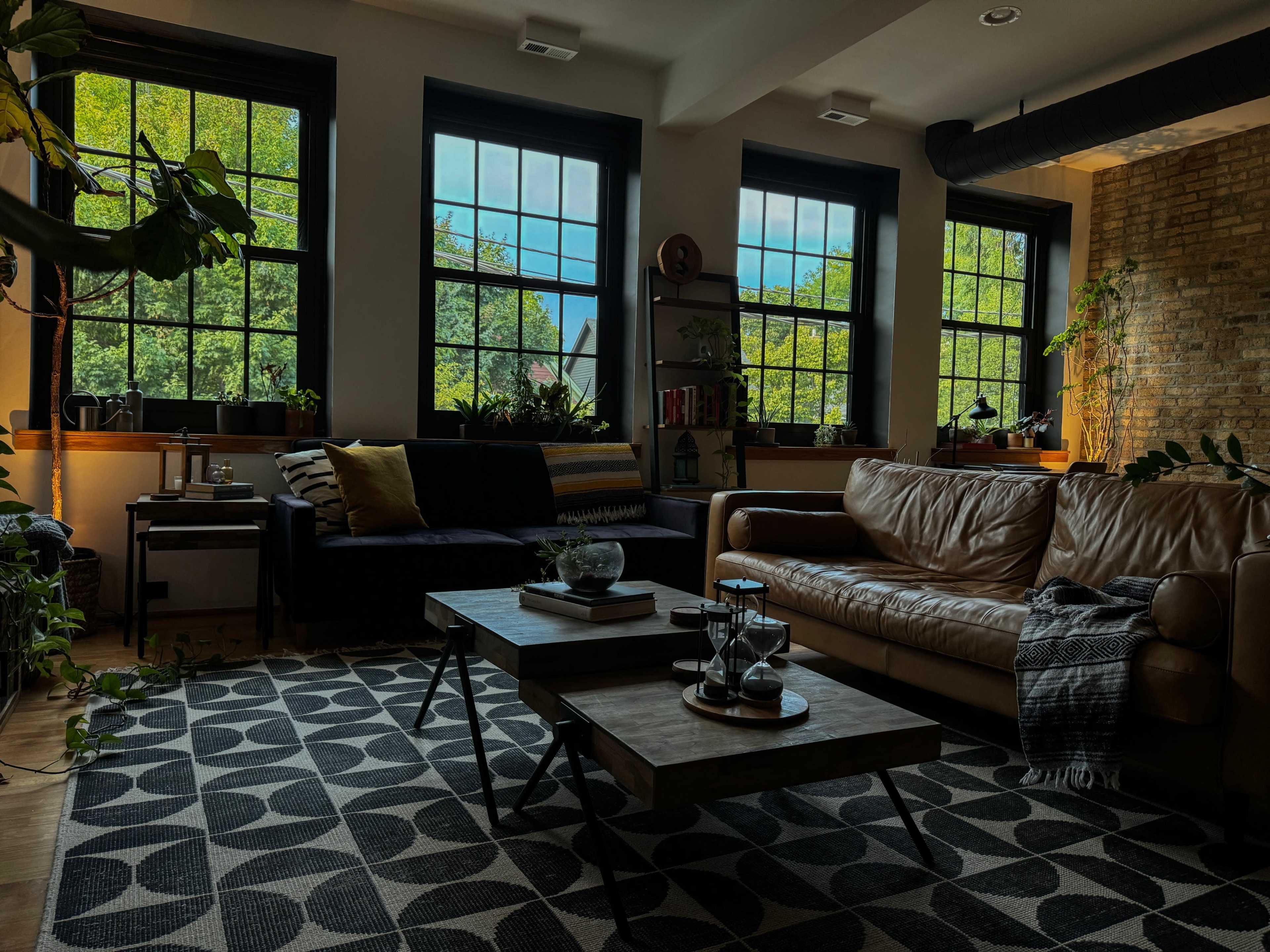 A cozy living room features large windows with greenery outside, a brown leather sofa, a patterned rug, and wooden tables decorated with plants and a clear vase.