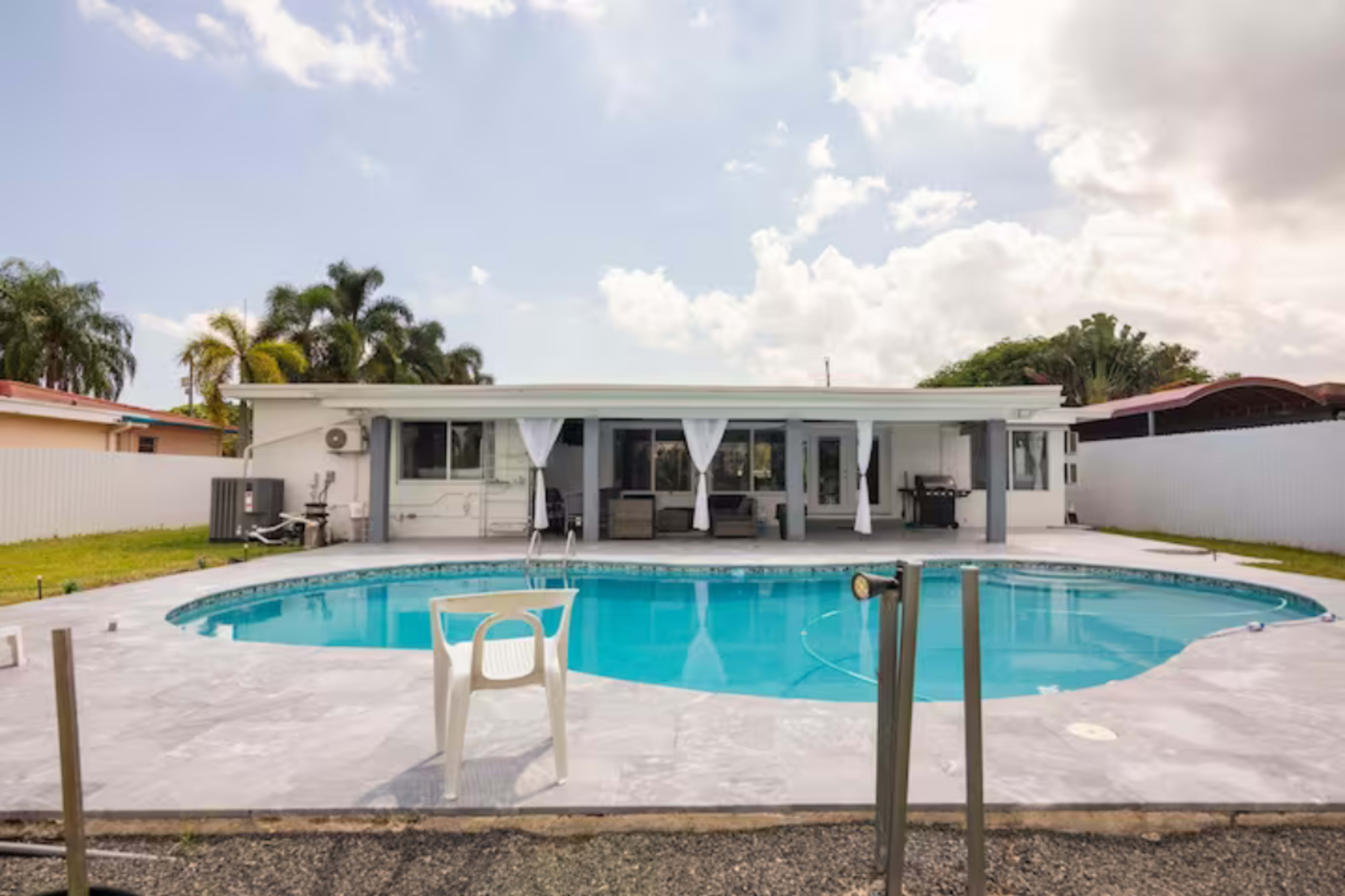 A modern single-story house with a swimming pool in the backyard, surrounded by palm trees and a grassy area.