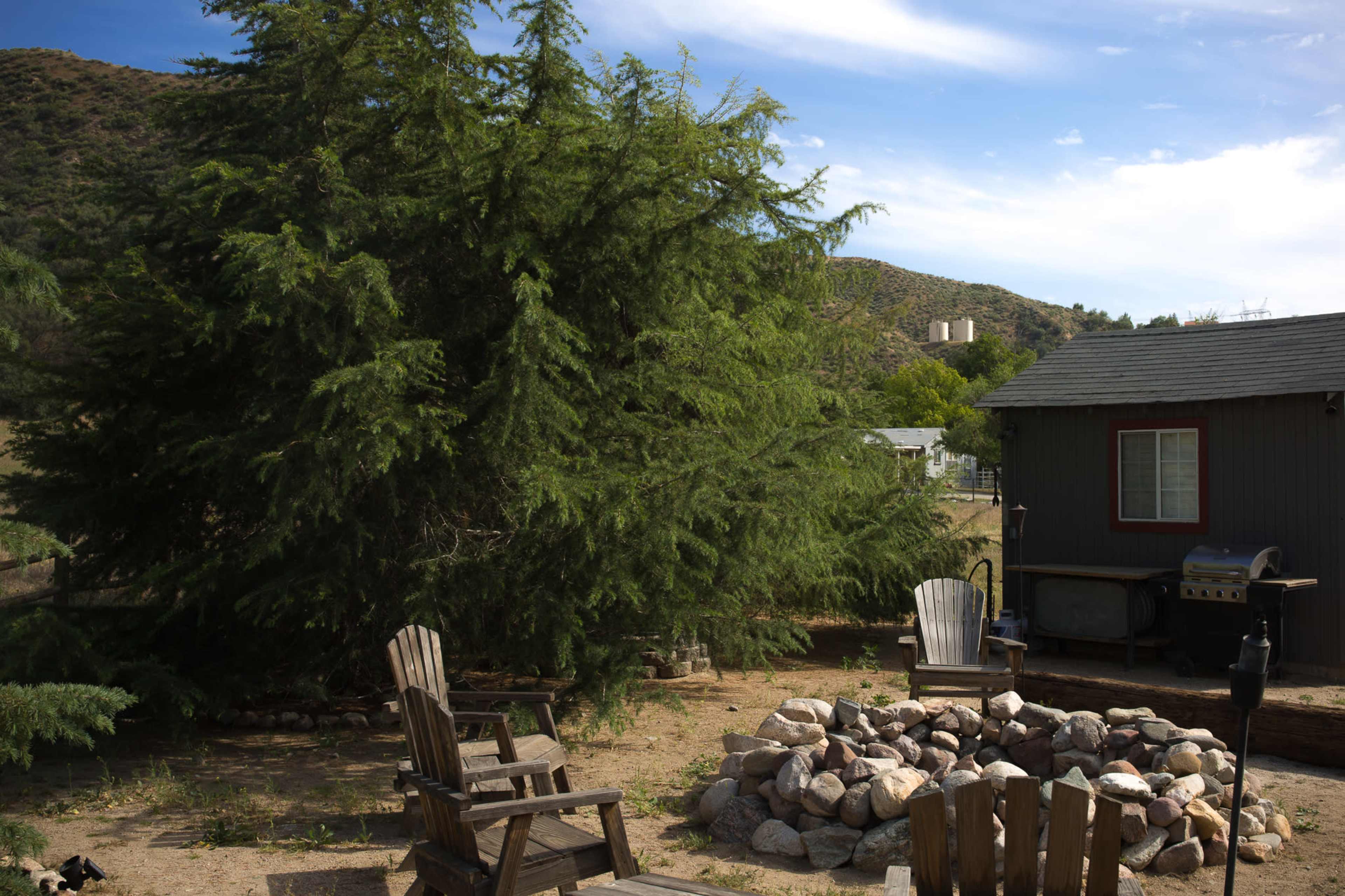 The image shows a rustic outdoor setting with a stone fire pit surrounded by wooden chairs, near a small gray house and large trees, against a hilly background.