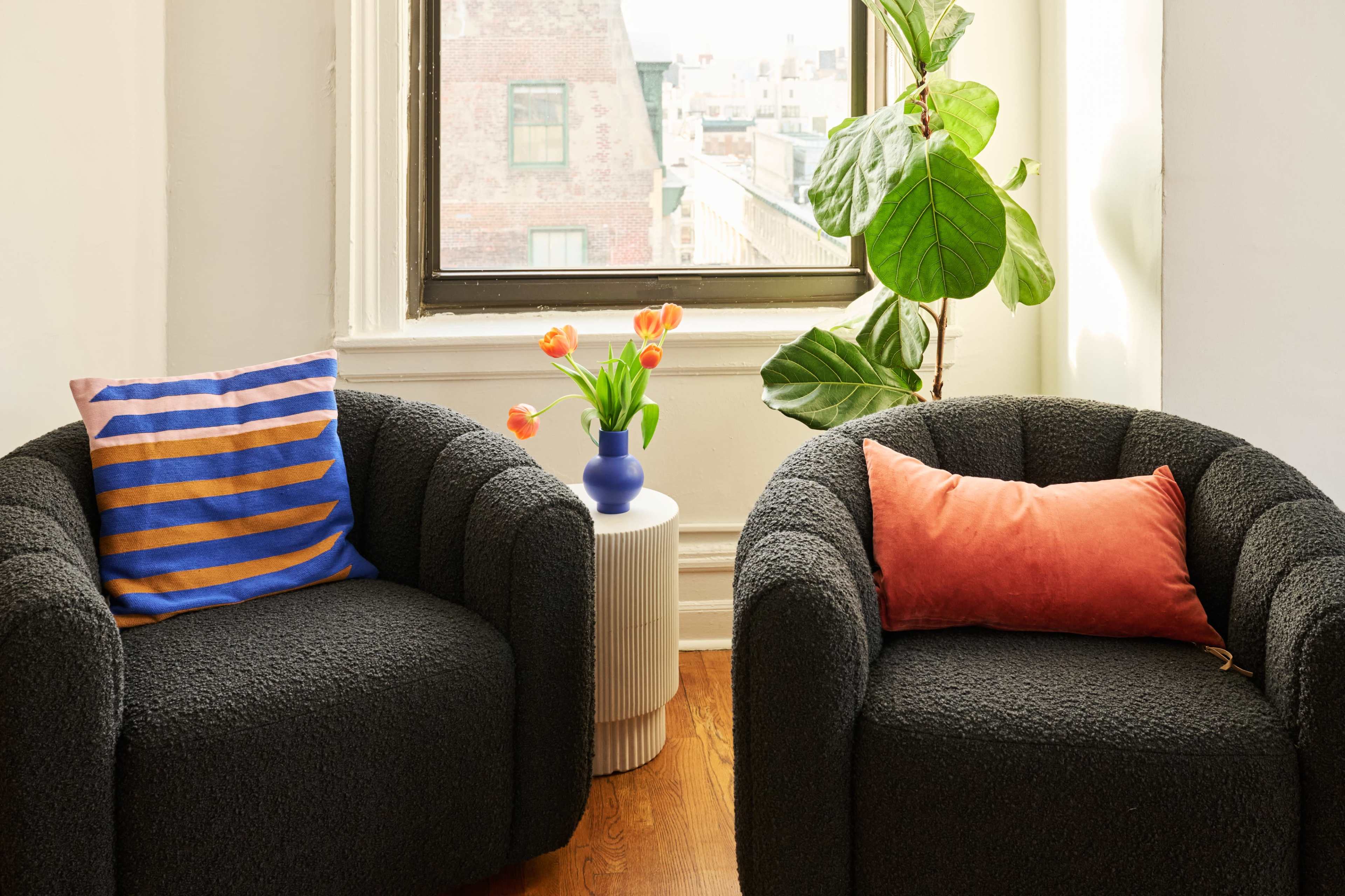A cozy seating area features two textured black chairs flanking a small white side table with a blue vase holding orange tulips, beside a large leafy plant and a window overlooking a cityscape.